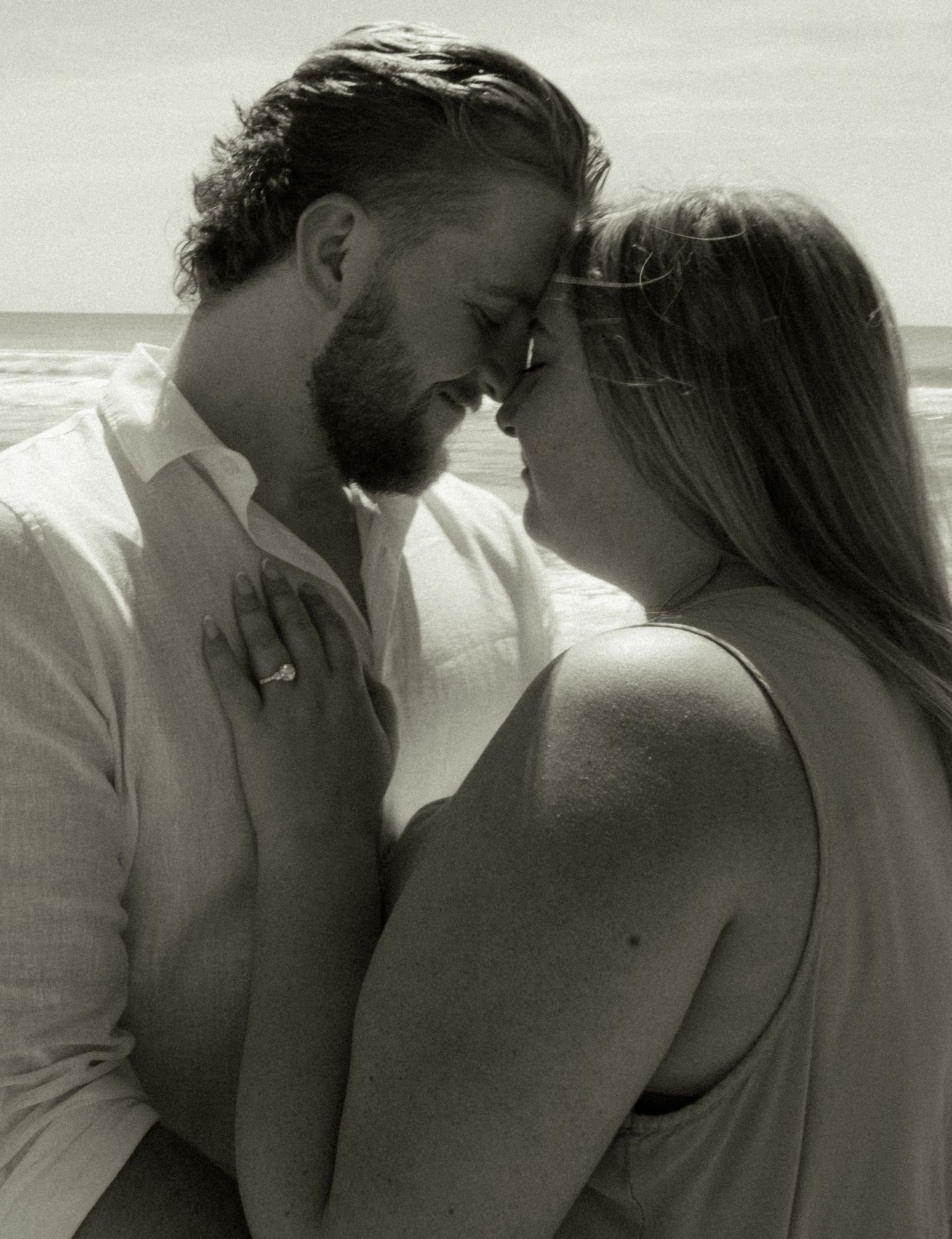 Black and white beach photo of a couple sharing a romantic moment, woman wearing an engagement ring.