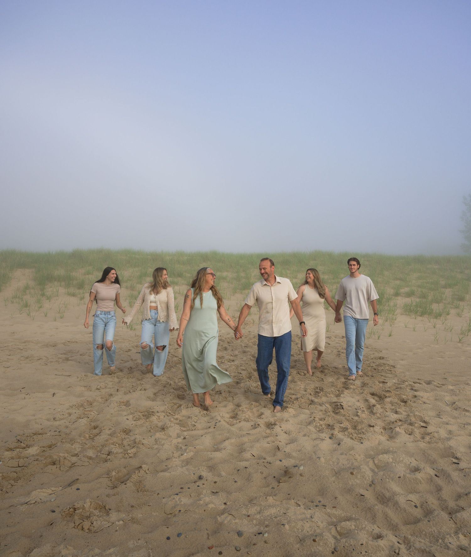 A joyful family of seven walks hand-in-hand on a sandy beach under a misty sky.