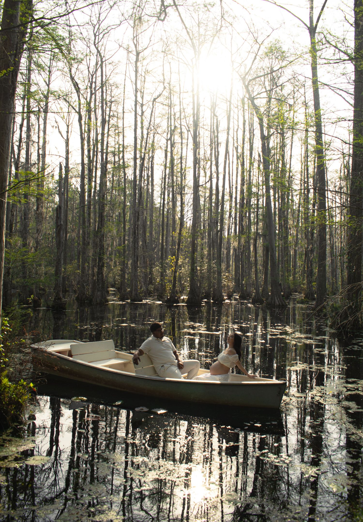 Couple sitting in a small boat on a serene swamp surrounded by tall trees with sunlight filtering through.
