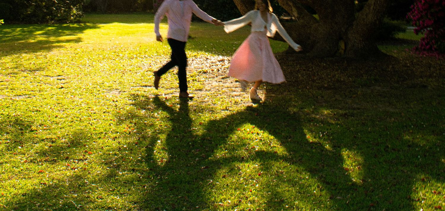 Couple holding hands running on lush green grass in golden sunlight near a tree, casting long shadows.