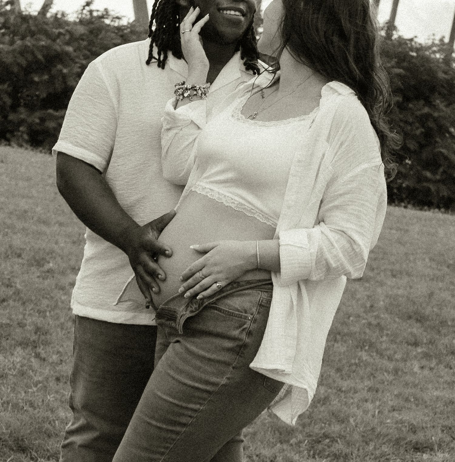 Black and white maternity photo of a couple outdoors, man embracing pregnant woman from behind, both smiling.