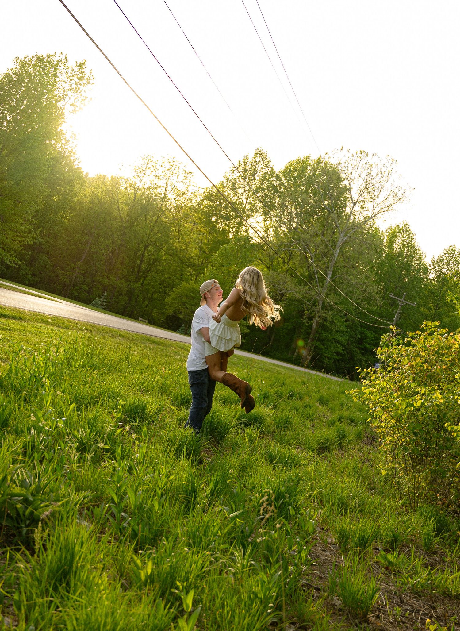 A couple joyfully spinning together in a sunlit green meadow surrounded by lush trees during golden hour.