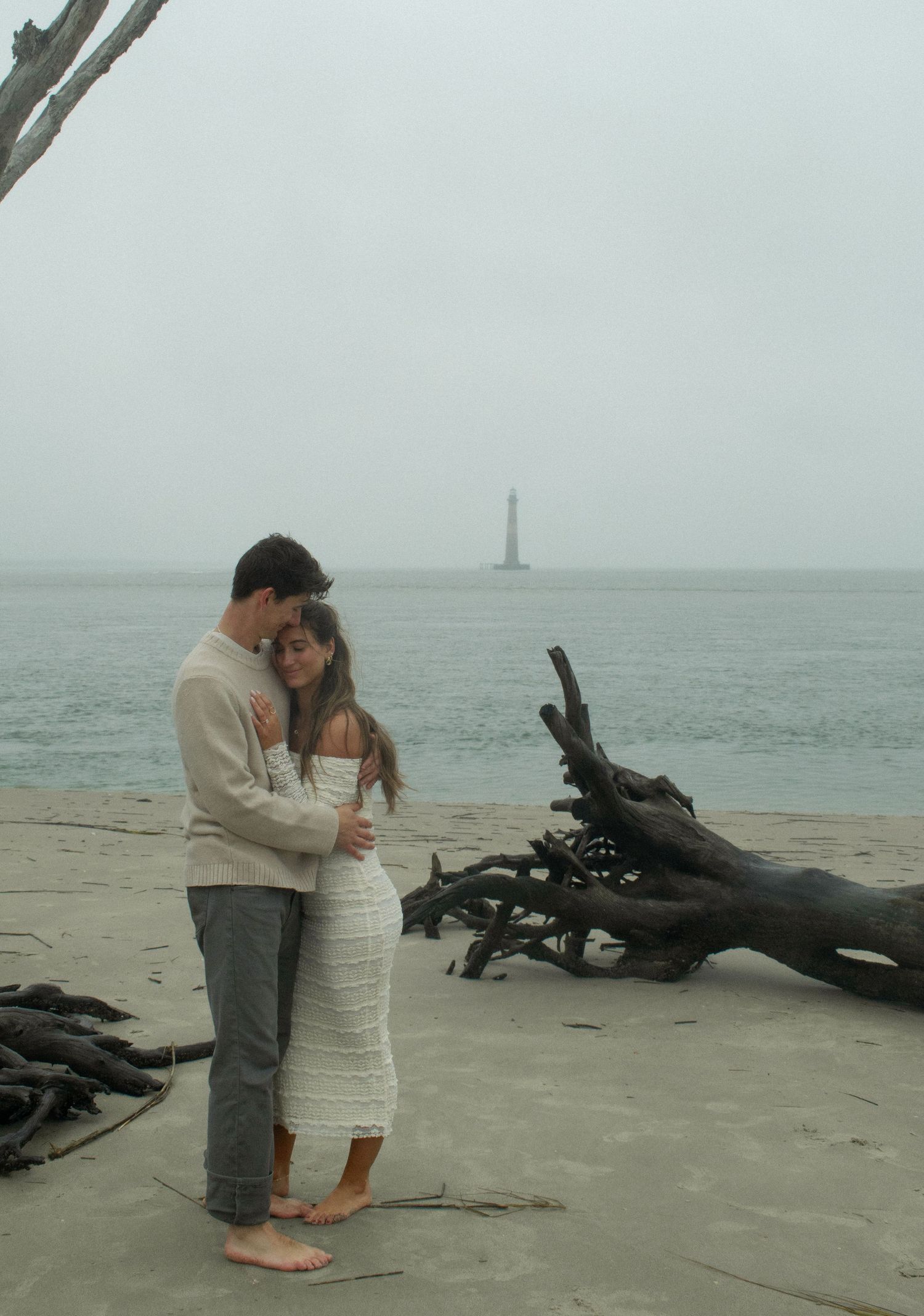 A couple shares a romantic moment on a beach with driftwood and a lighthouse in the background.