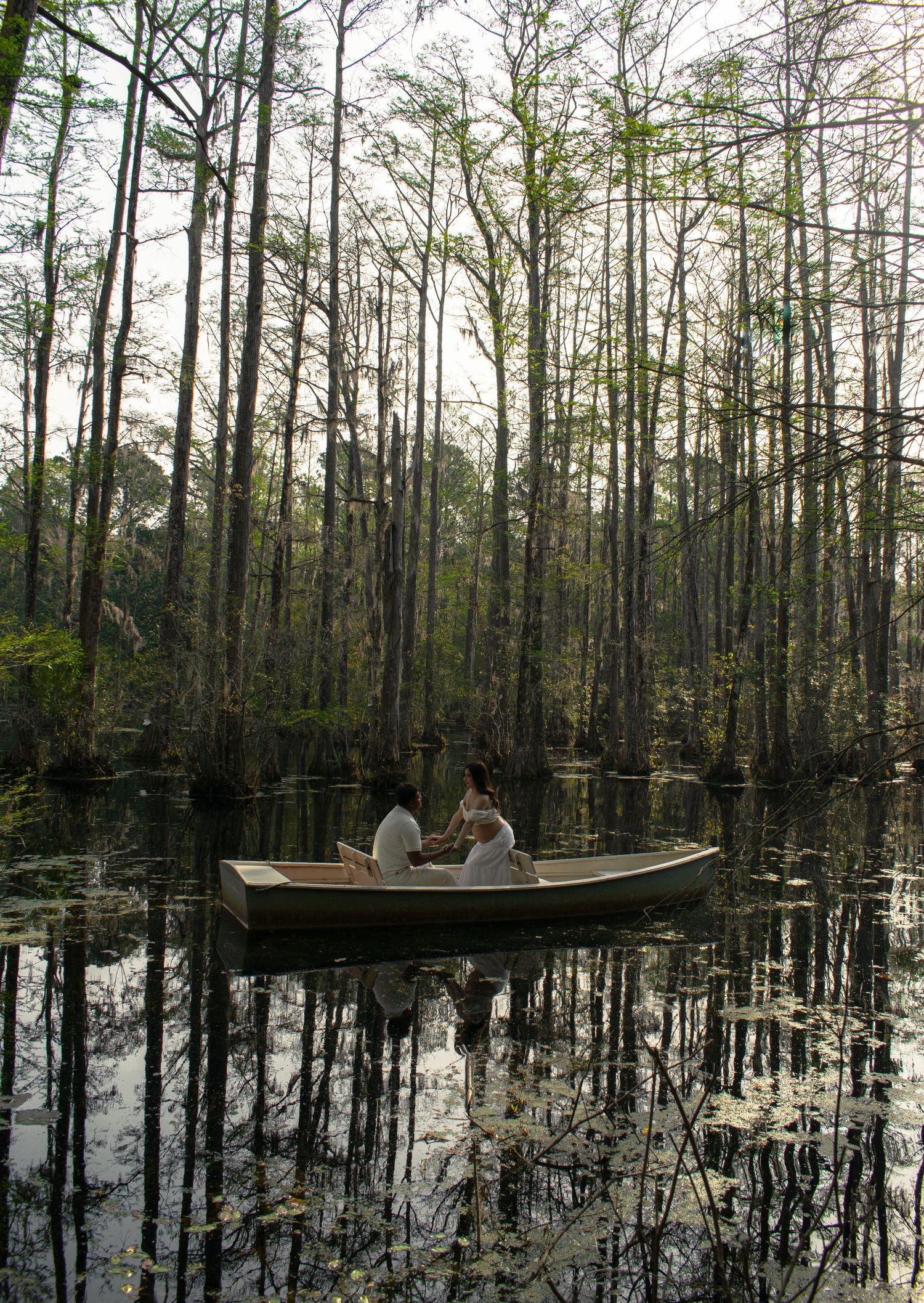 Two people sitting in a rowboat on a calm swamp surrounded by tall cypress trees and their reflections in still water.