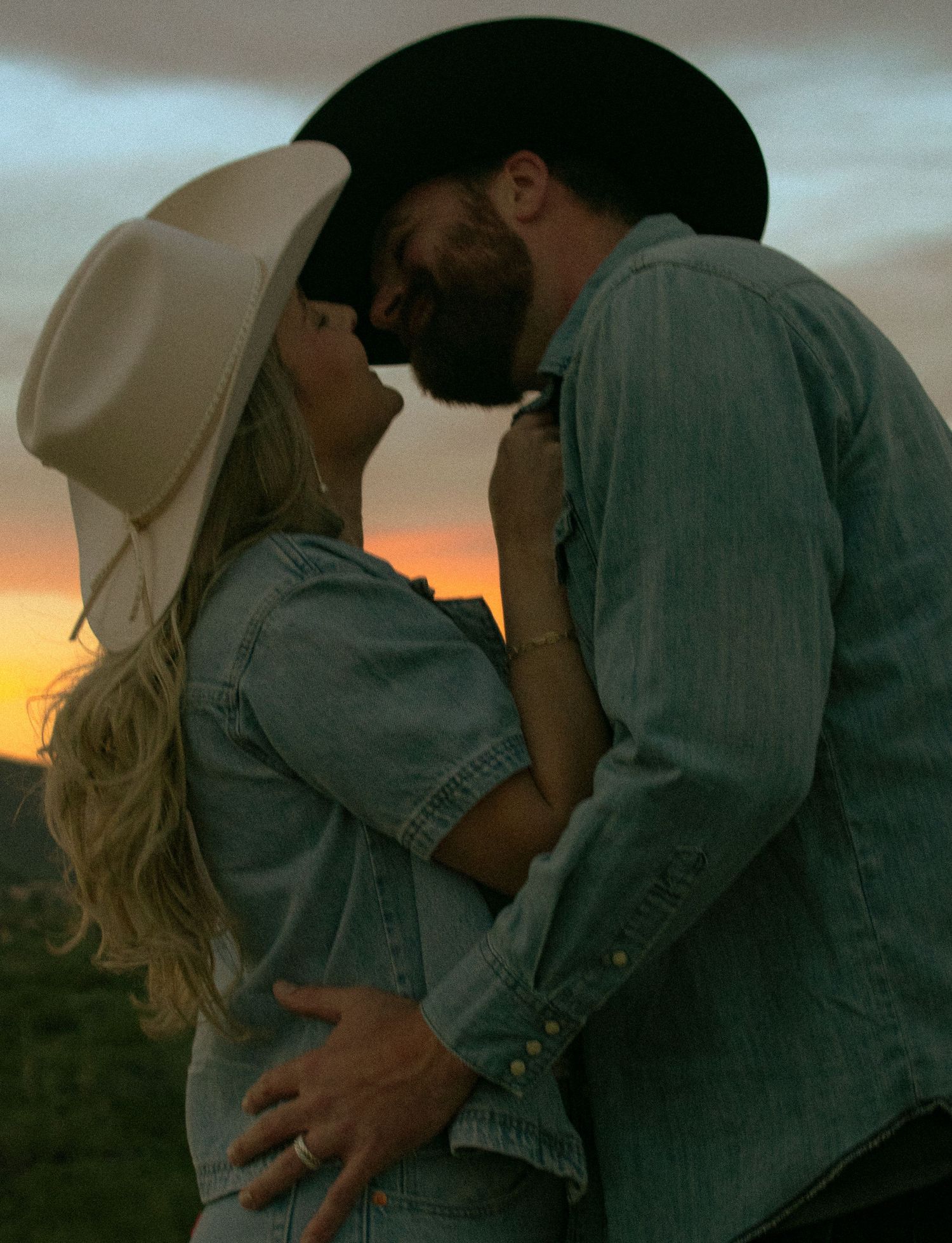 Couple in cowboy hats sharing a romantic kiss at sunset, wearing matching denim outfits in a golden outdoor setting.