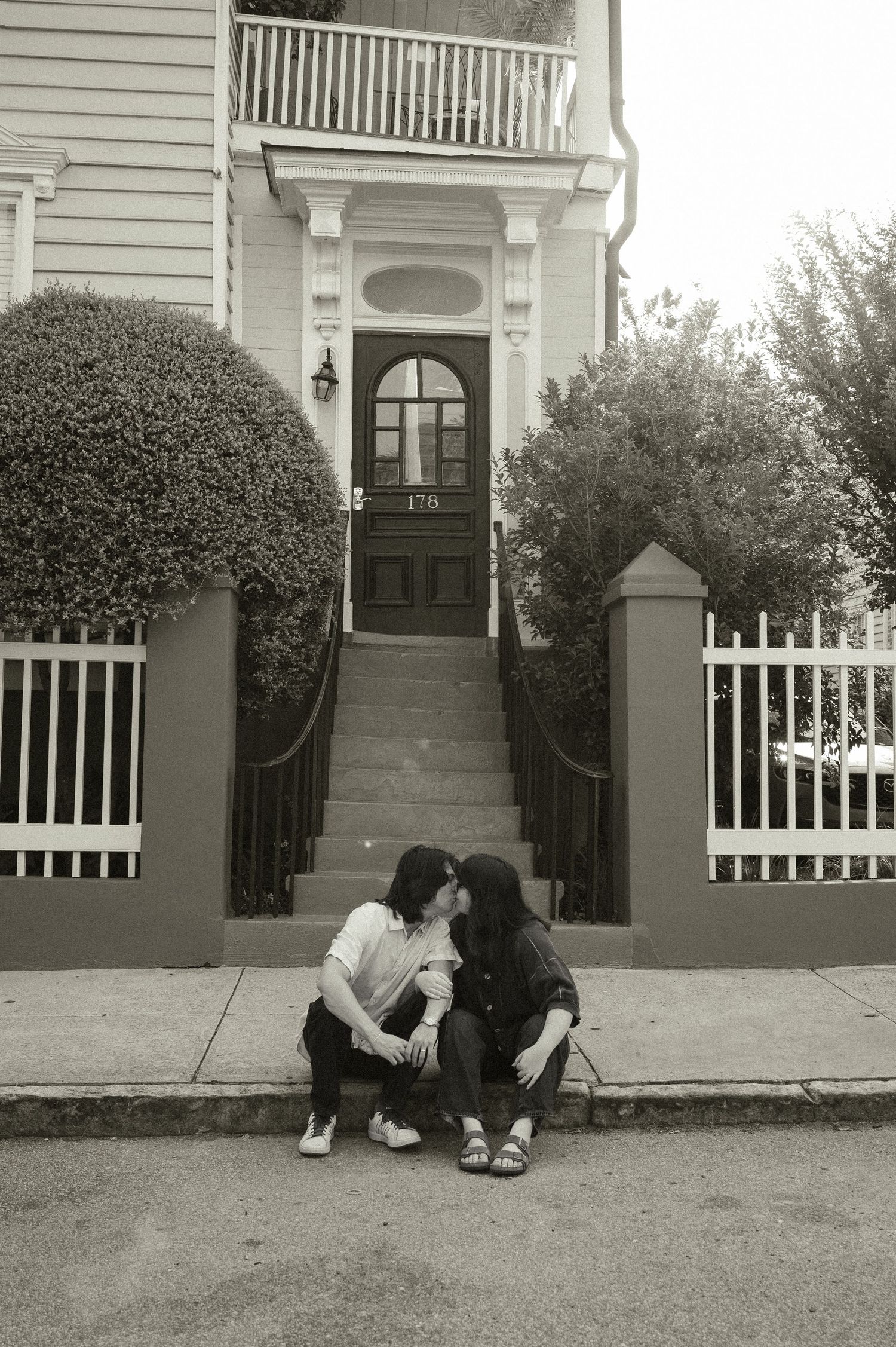 Black and white photo of a couple kissing while sitting on steps outside a classic Victorian home.