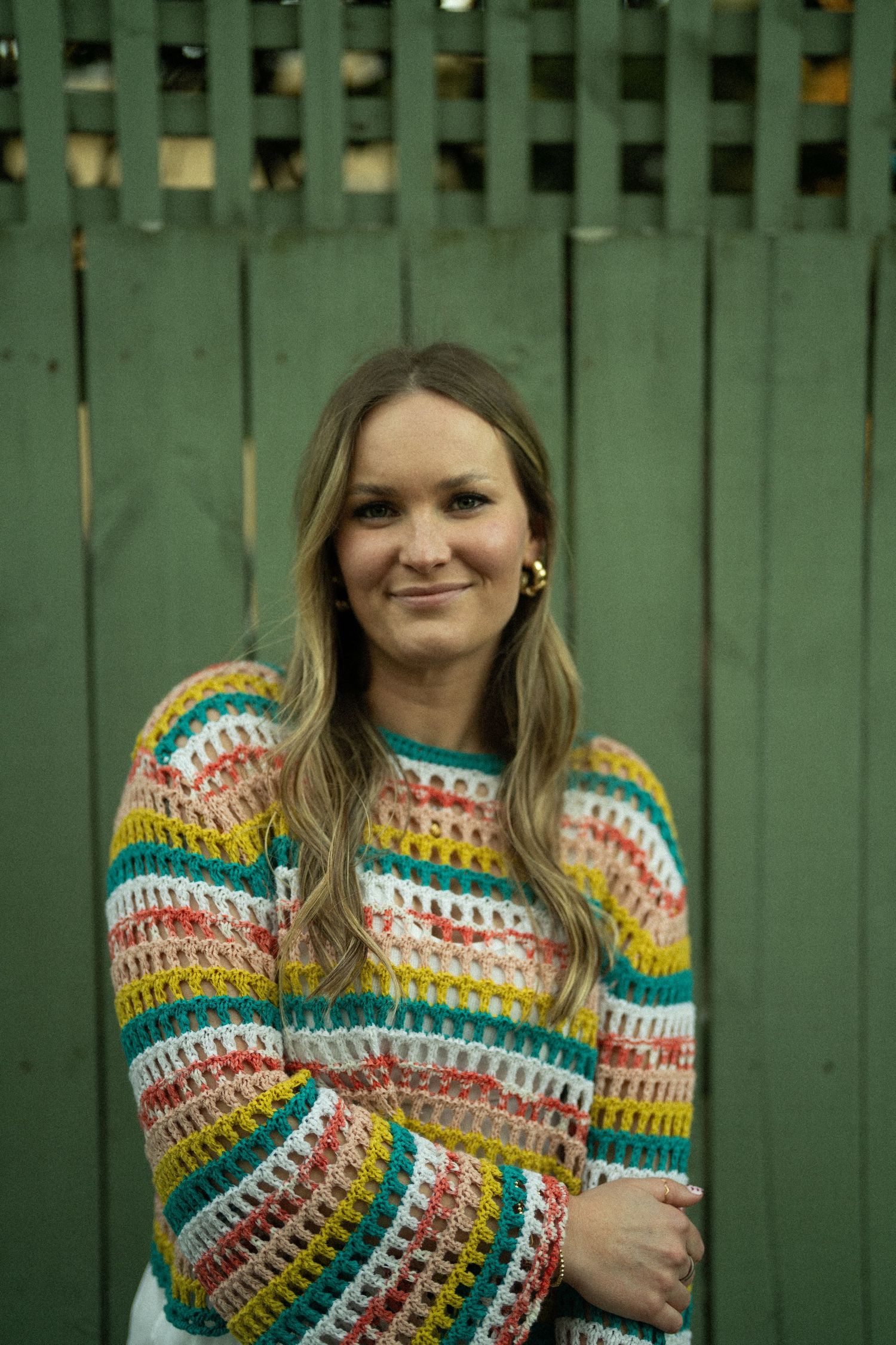 Woman smiling in colorful striped crochet sweater standing against a green wooden fence.