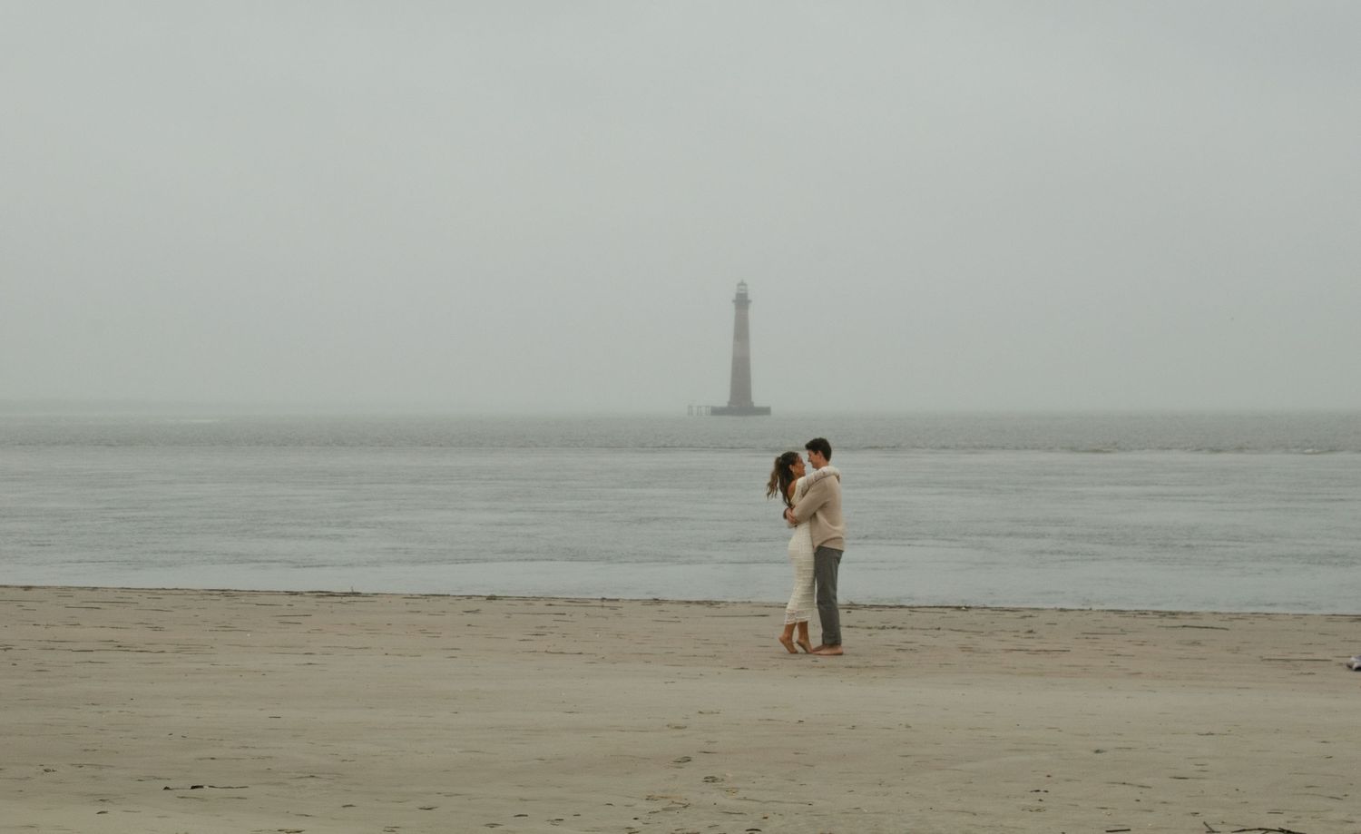 A couple embracing on a foggy beach with a lighthouse in the distance.