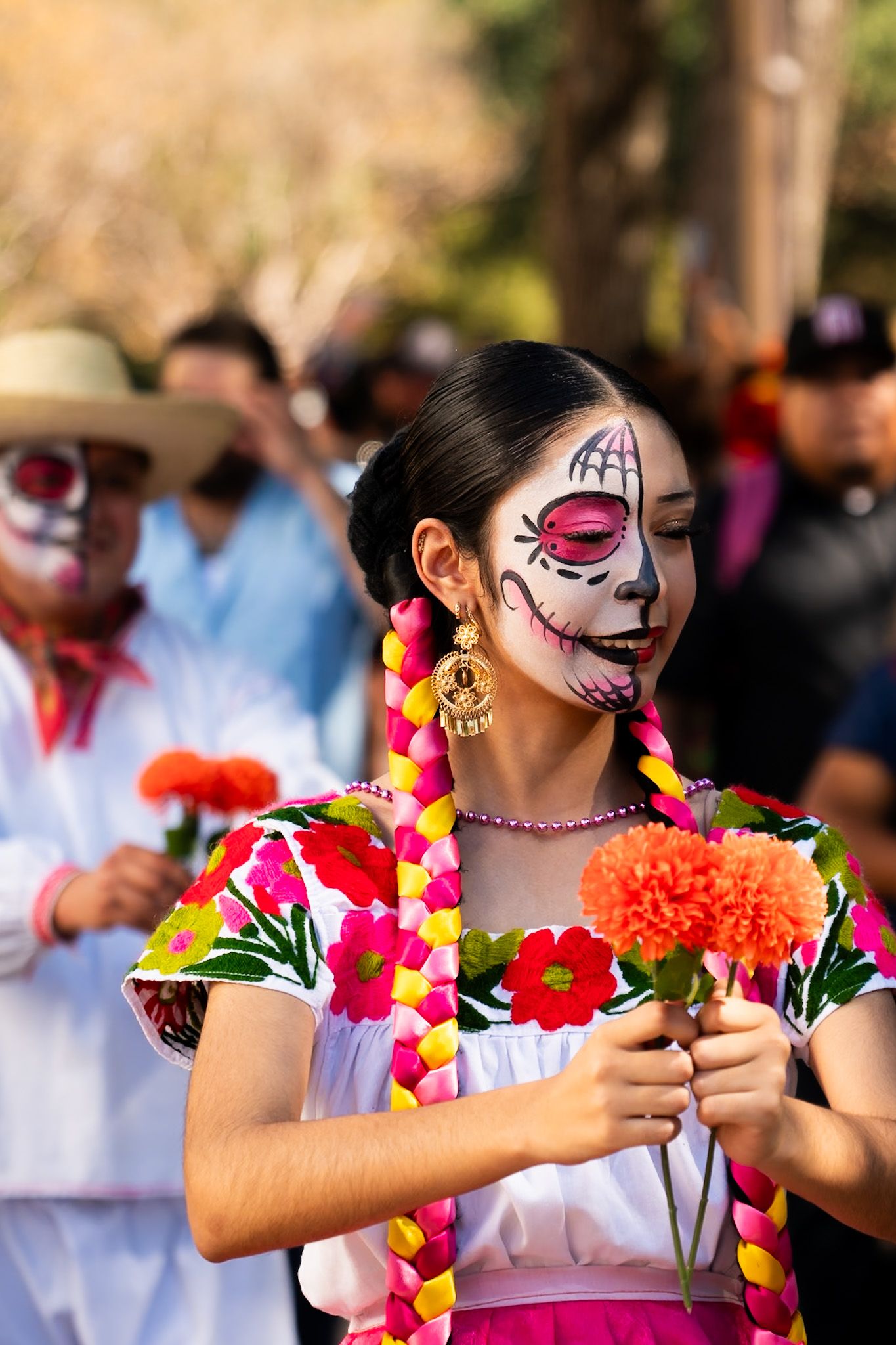 Performers in white dresses with face paint and floral leis dance at an outdoor festival.