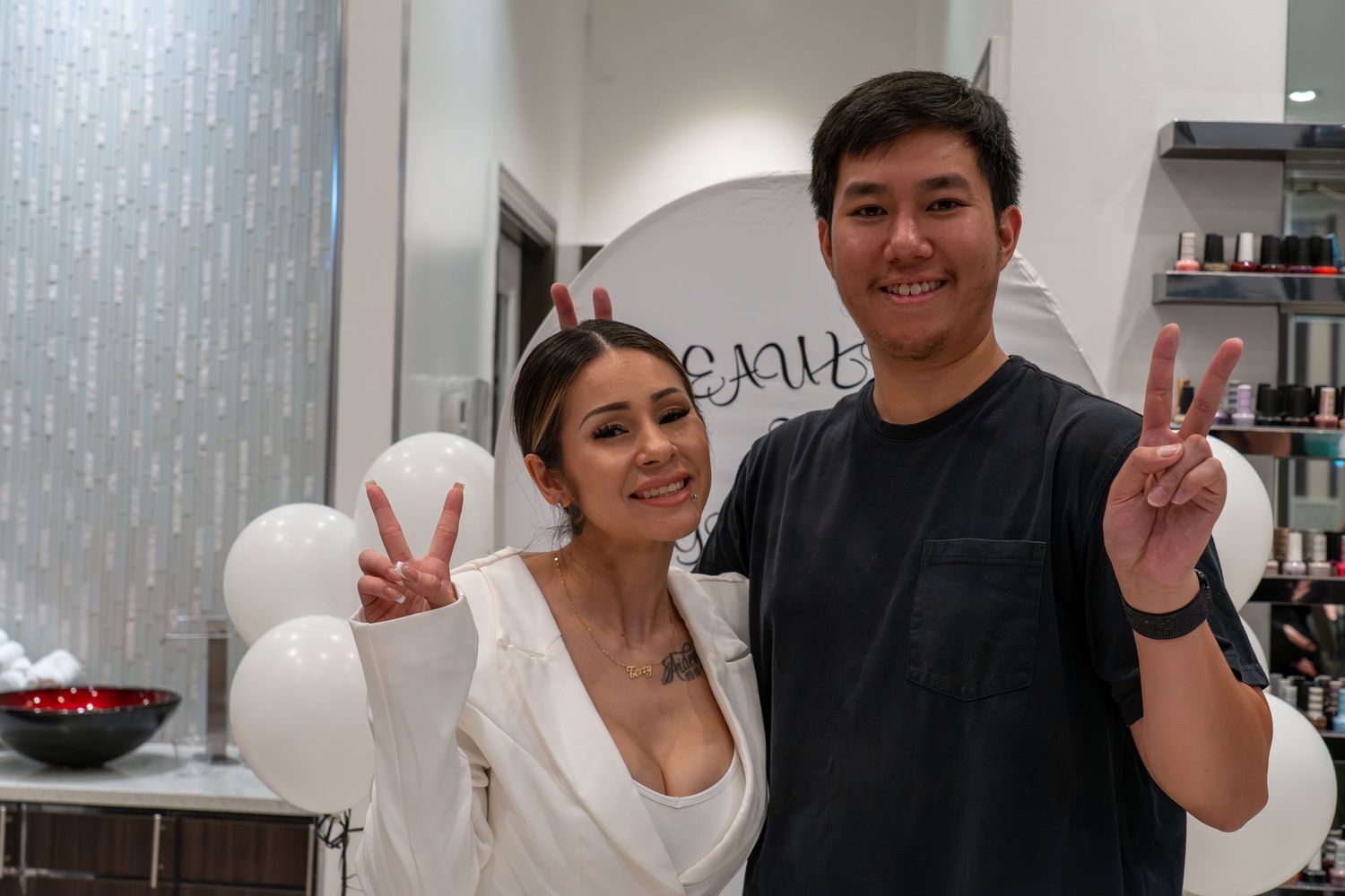 Two people pose together in a salon setting with a white balloon and beauty product shelves in the background.