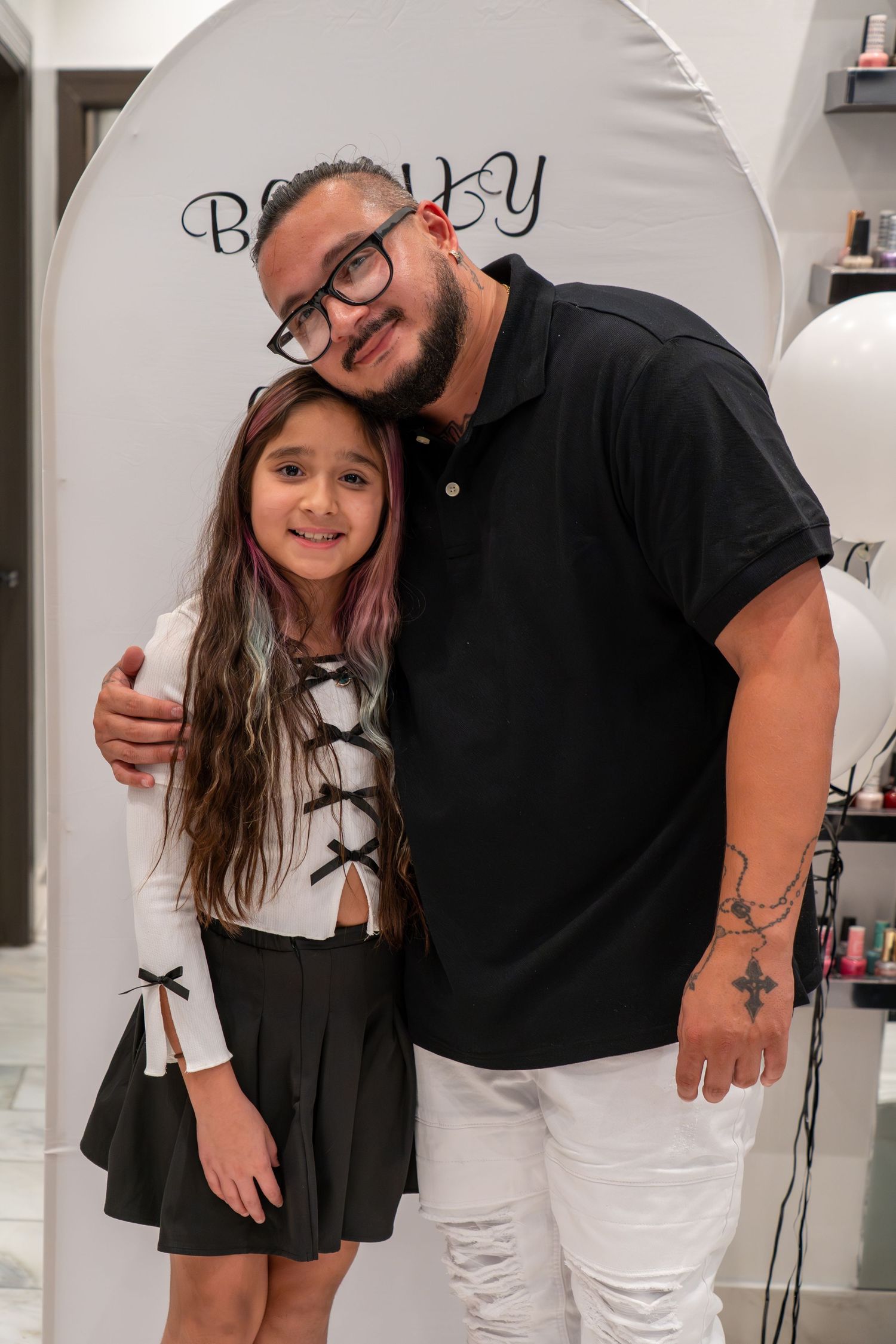 Two people pose together against a white wall with 'Beauty' text visible in a salon setting.