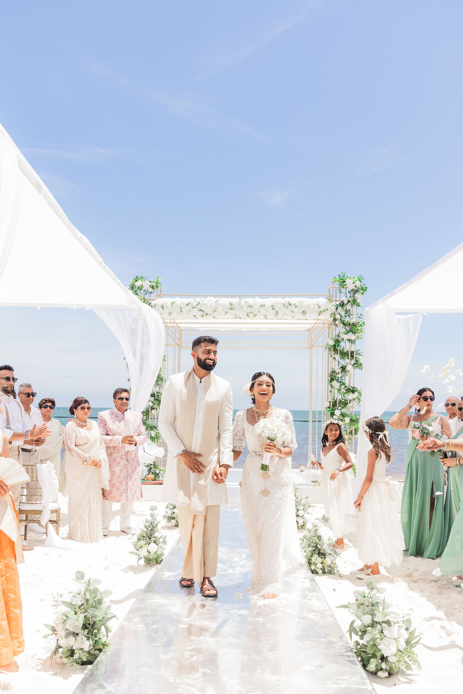 Couple in white attire walks down aisle at elegant outdoor beach wedding ceremony with floral arch.