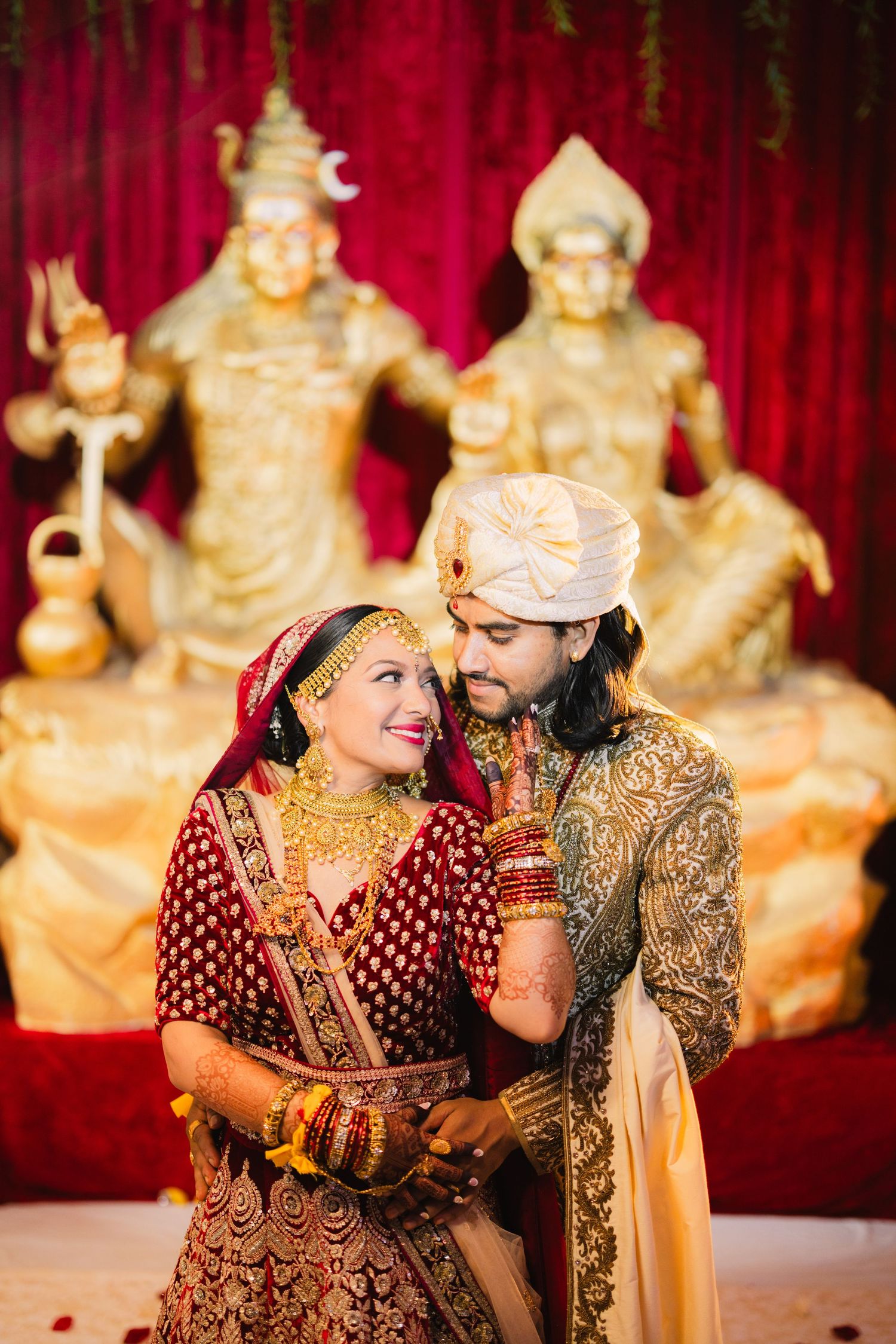 Indian bride in red lehenga and groom in sherwani pose before golden deity statues at traditional Hindu wedding ceremony.