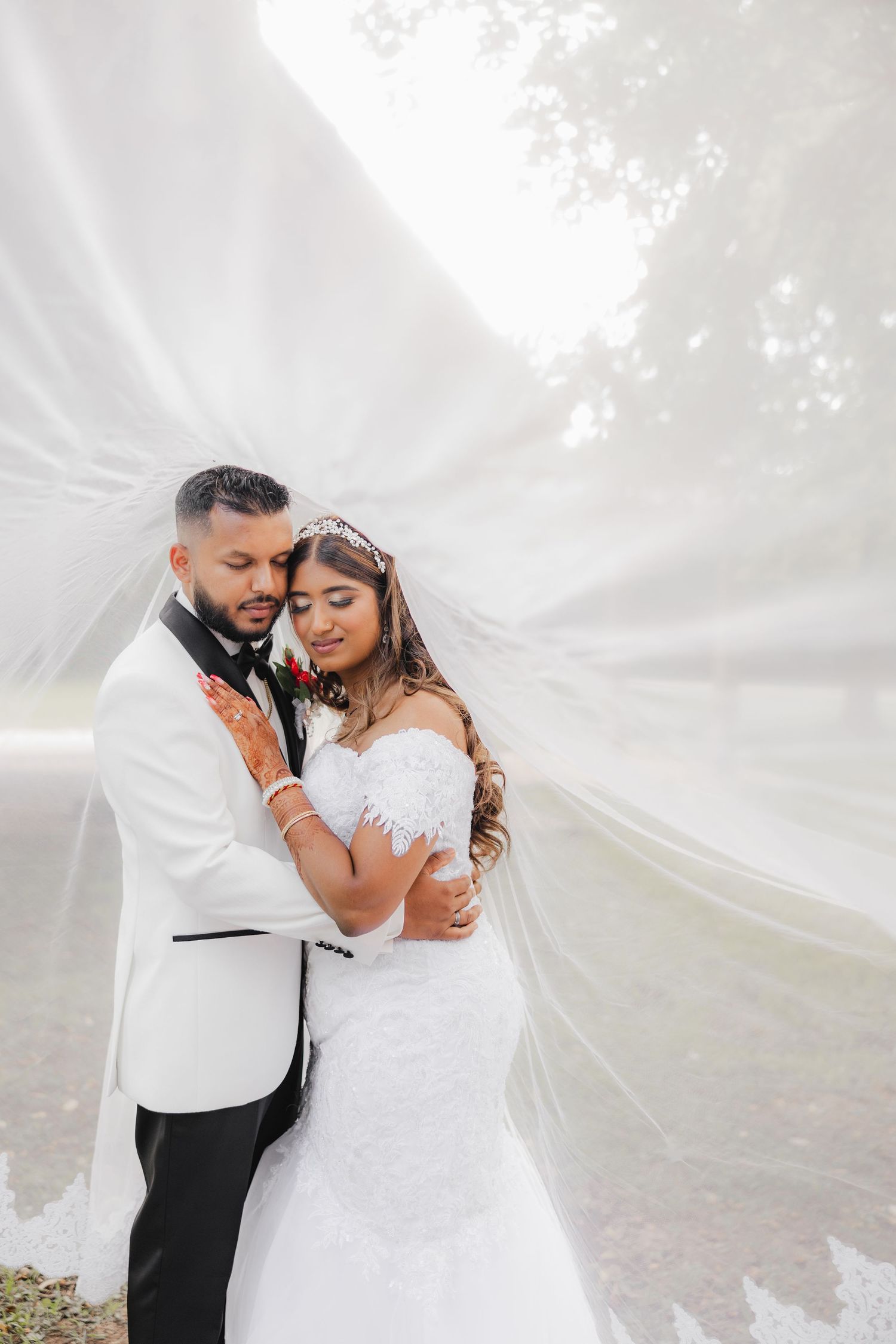 Bride and groom embrace under flowing white veil outdoors, bride in lace gown with tiara, groom in white tuxedo jacket.