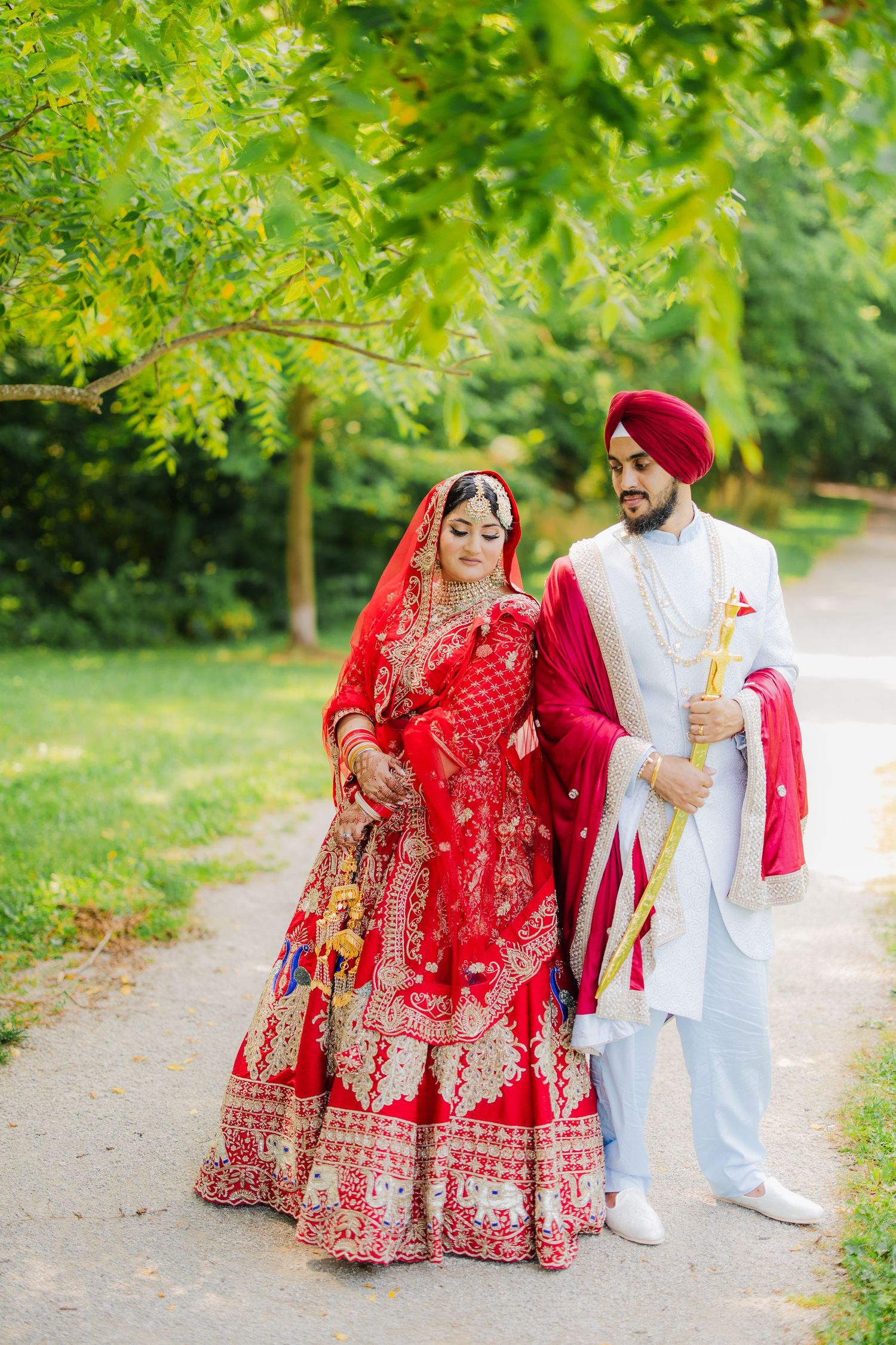 Sikh bride in red lehenga and groom in white sherwani with turban walk together on a garden path.