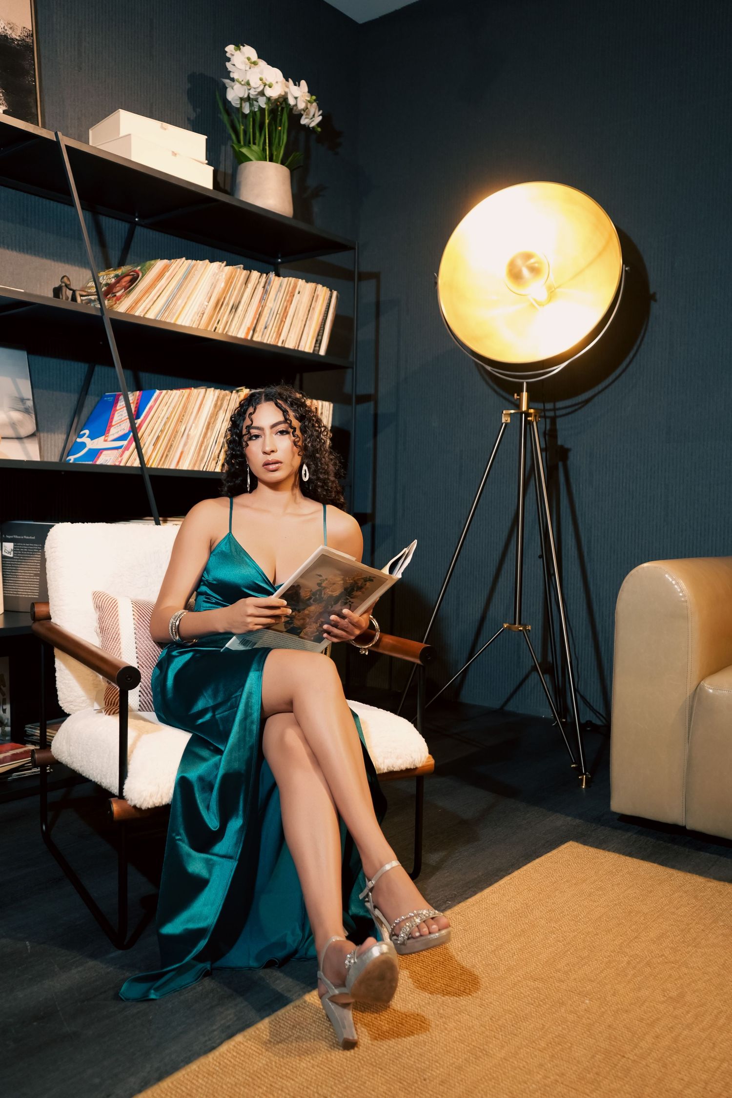 A person in an emerald green dress sits in a studio setting with dramatic lighting and vintage decor.