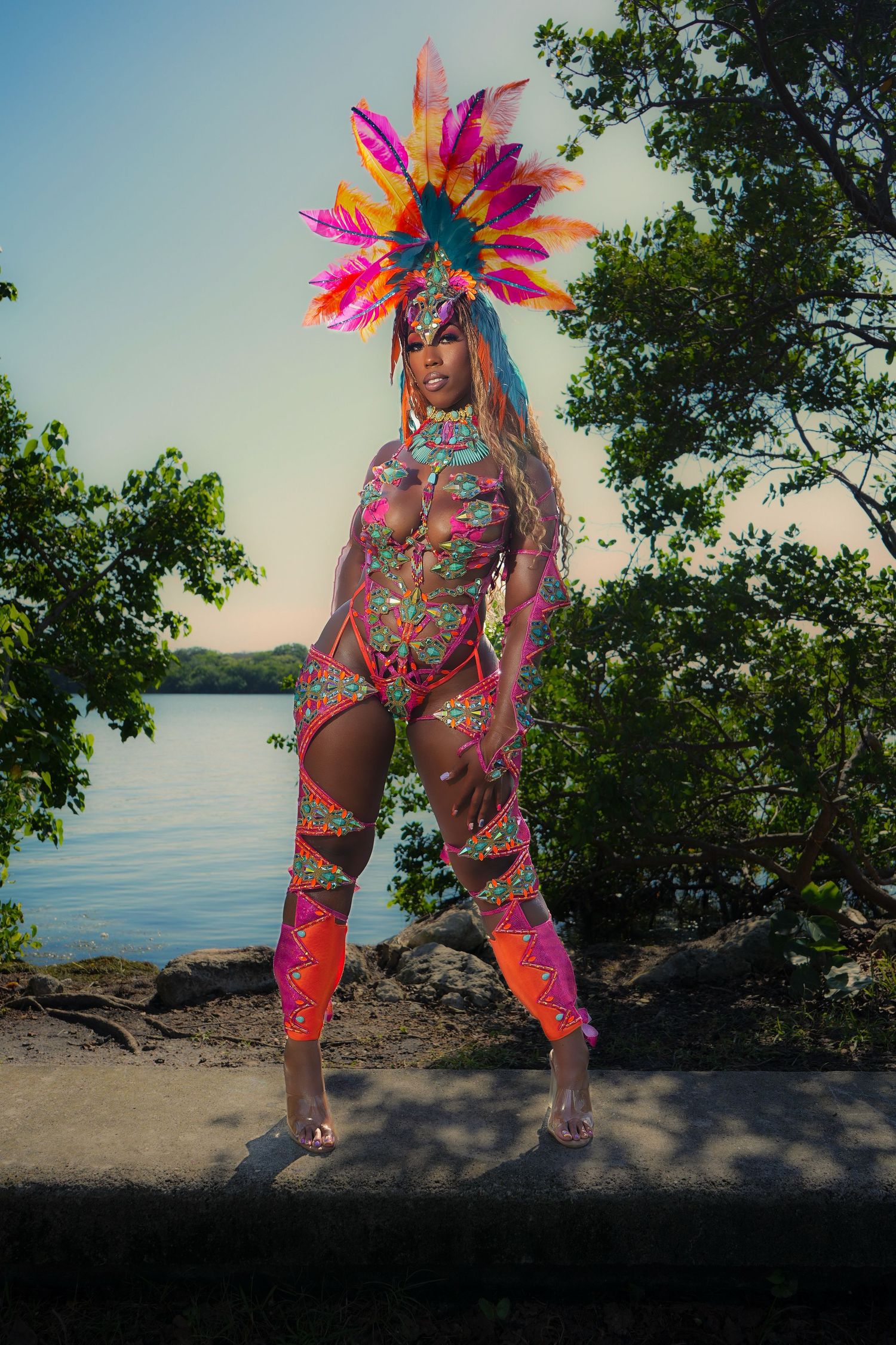 Vibrant carnival costume with pink and orange feathers photographed against a waterfront nature backdrop at sunset.