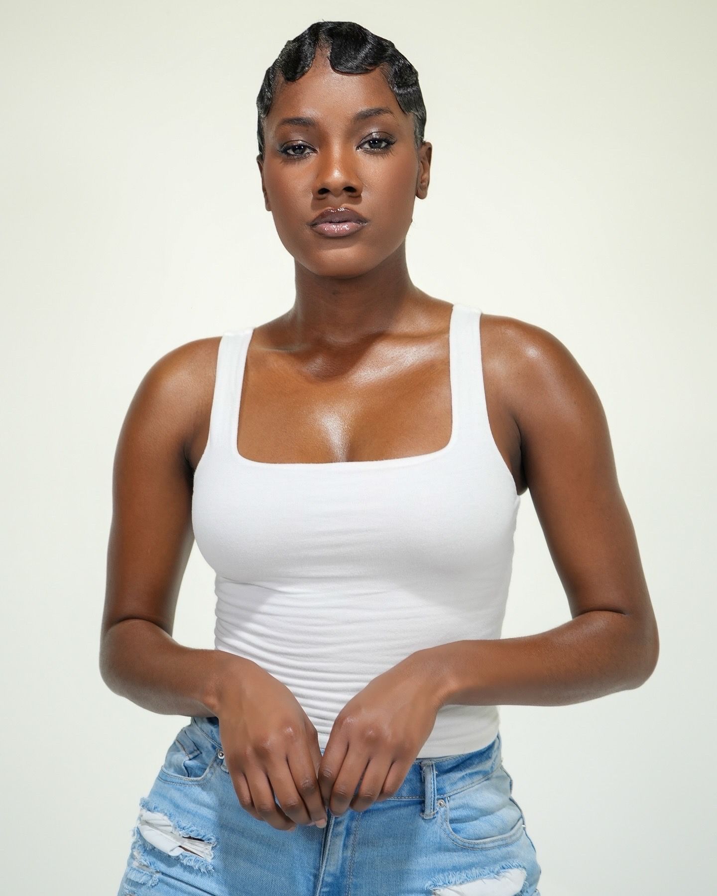 Model in white tank top and distressed denim jeans poses against light background with styled hair.