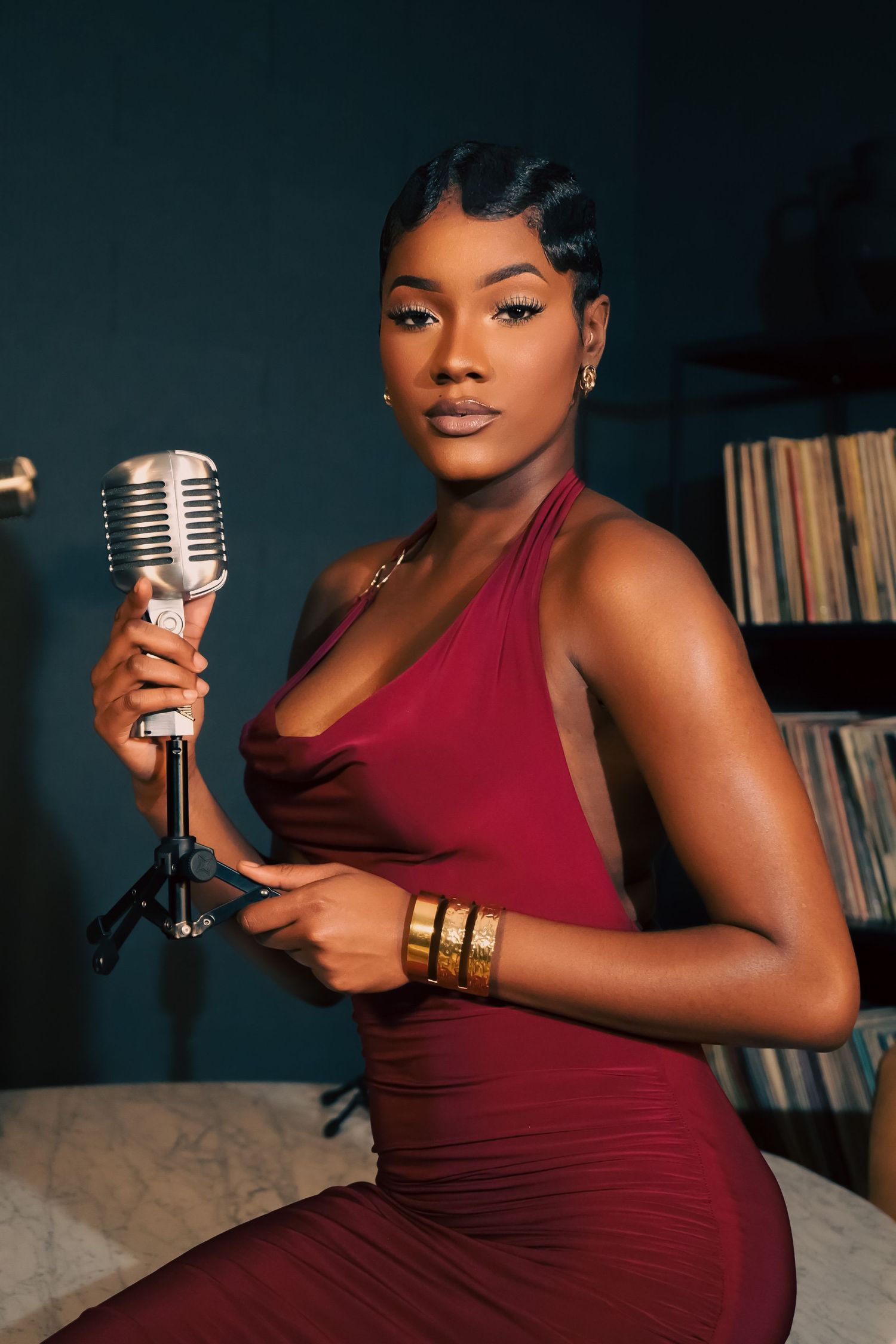 Singer in burgundy dress poses with vintage microphone against dark background with vinyl records.