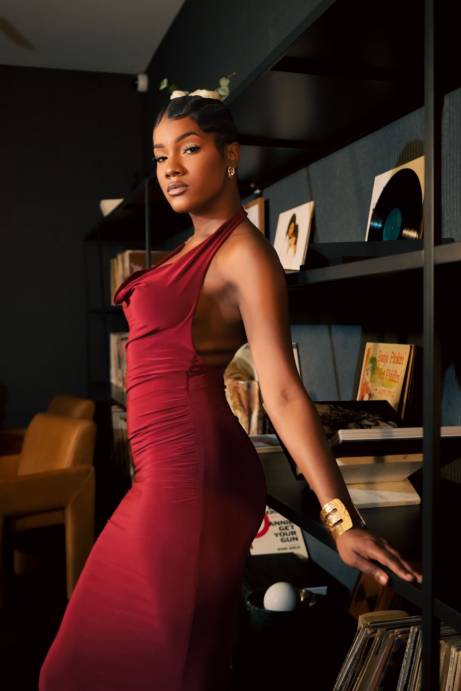 A person in a burgundy dress poses elegantly against a dark background with bookshelves visible behind them.