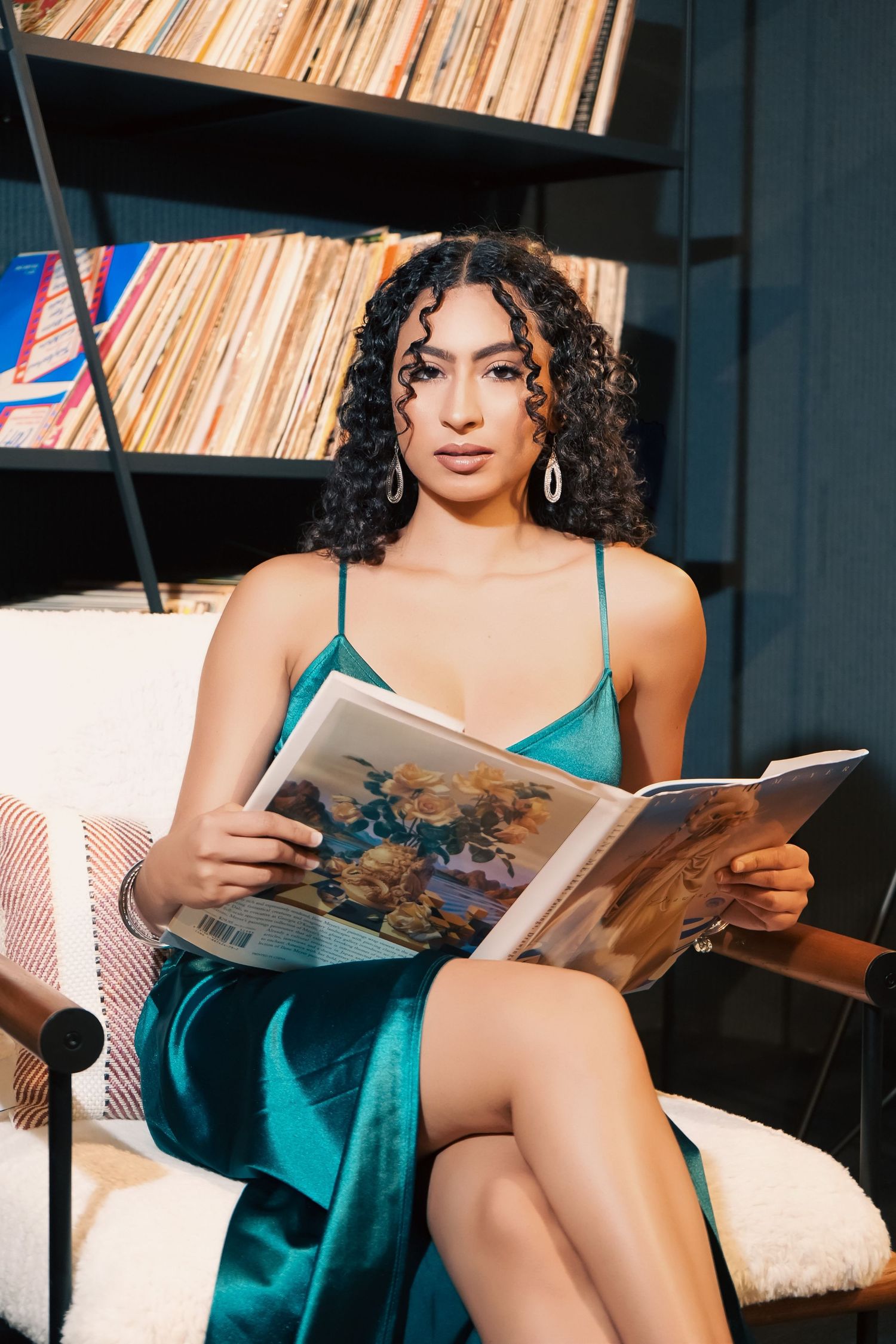 Woman in teal silk slip dress lounges on couch while reading magazine in moody lighting.