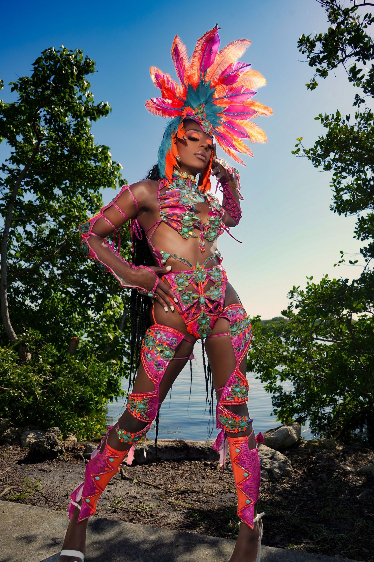 Carnival dancer in vibrant pink feathered costume poses dramatically against tropical beach landscape at sunset.