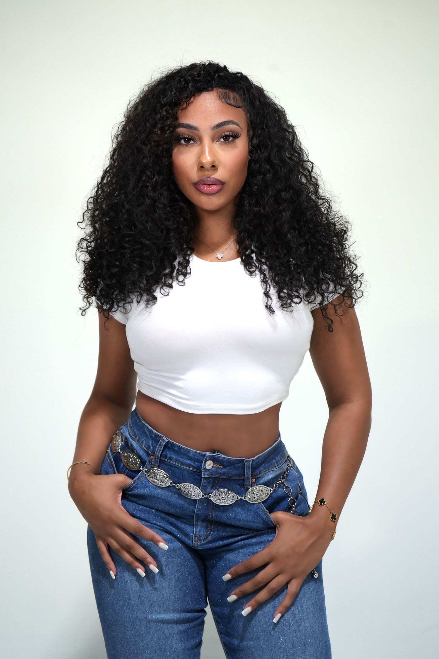 Fashion model wearing white crop top and blue jeans with curly natural hair against light background.