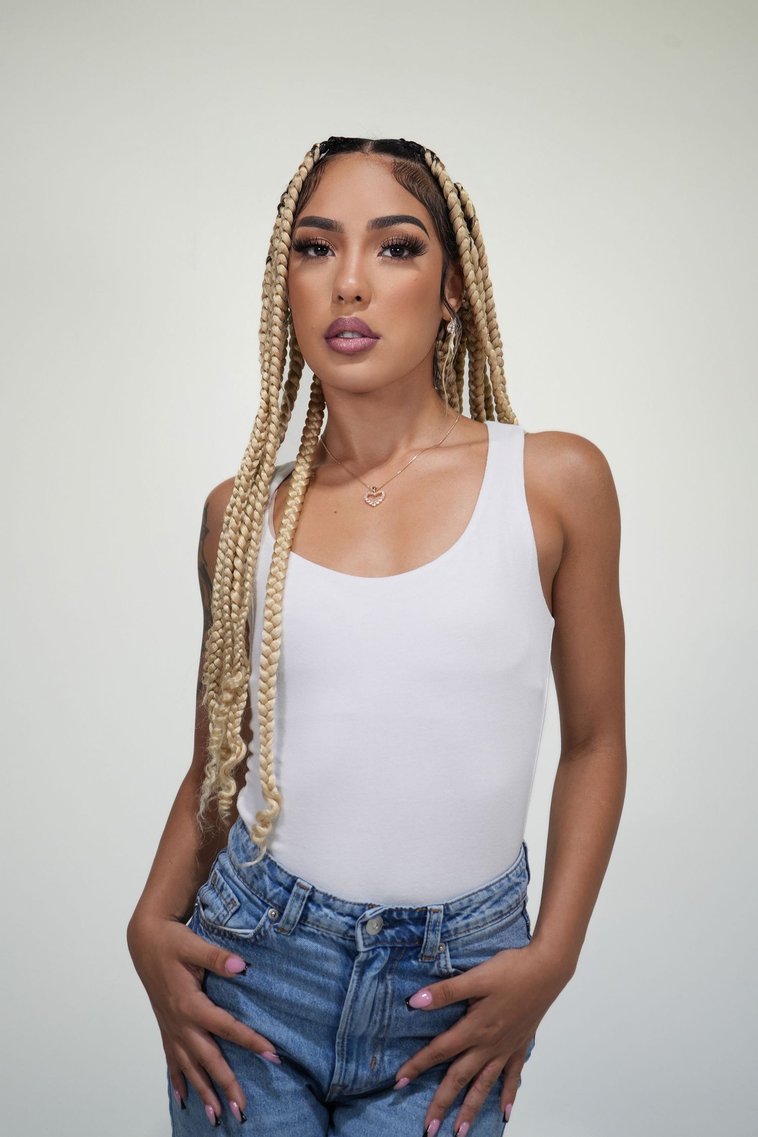Fashion model wearing white tank top and denim jeans poses against white background with blonde braided hairstyle.