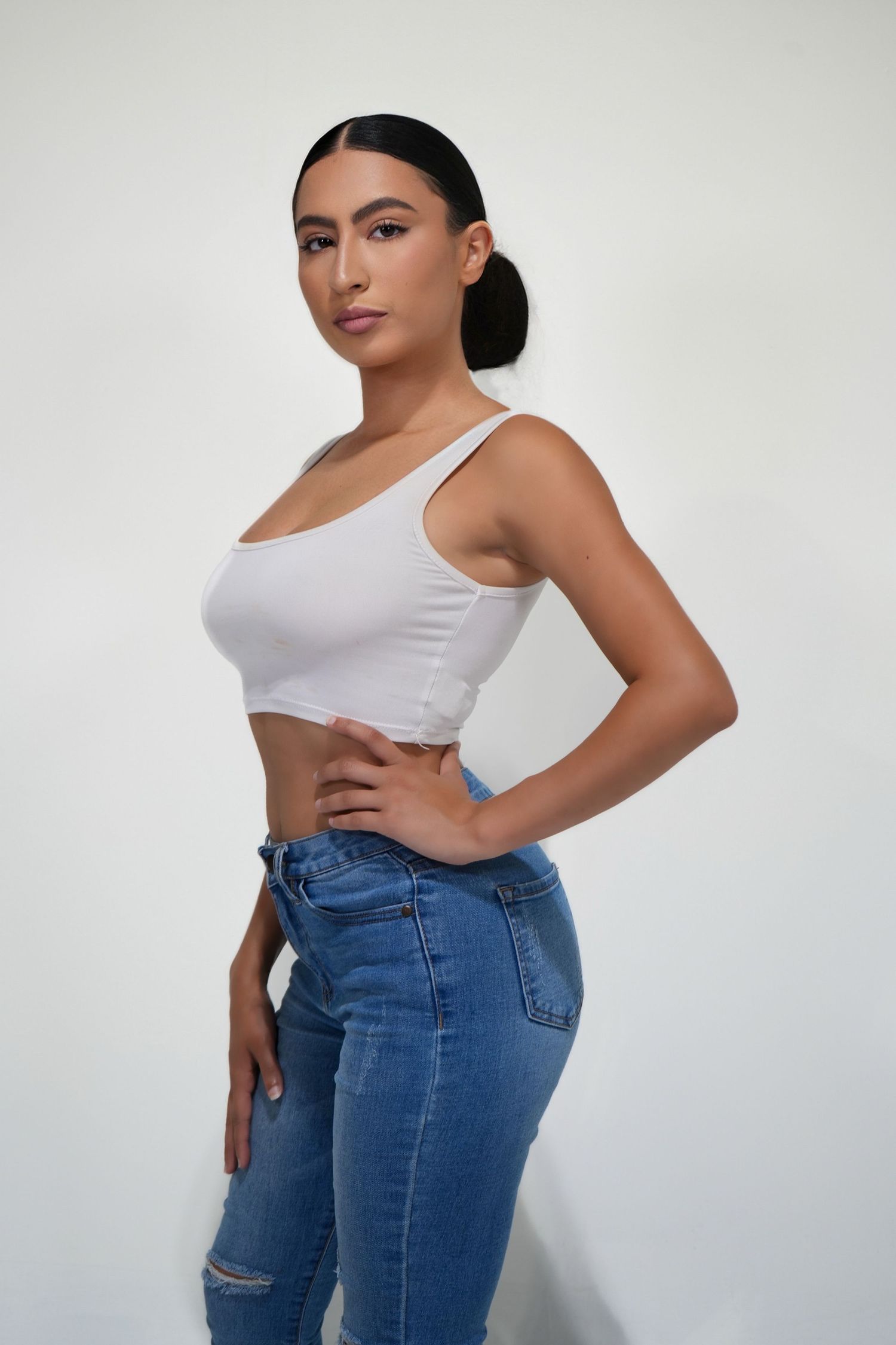 Model wearing white tank top and blue jeans poses against white background showing casual style and fit.