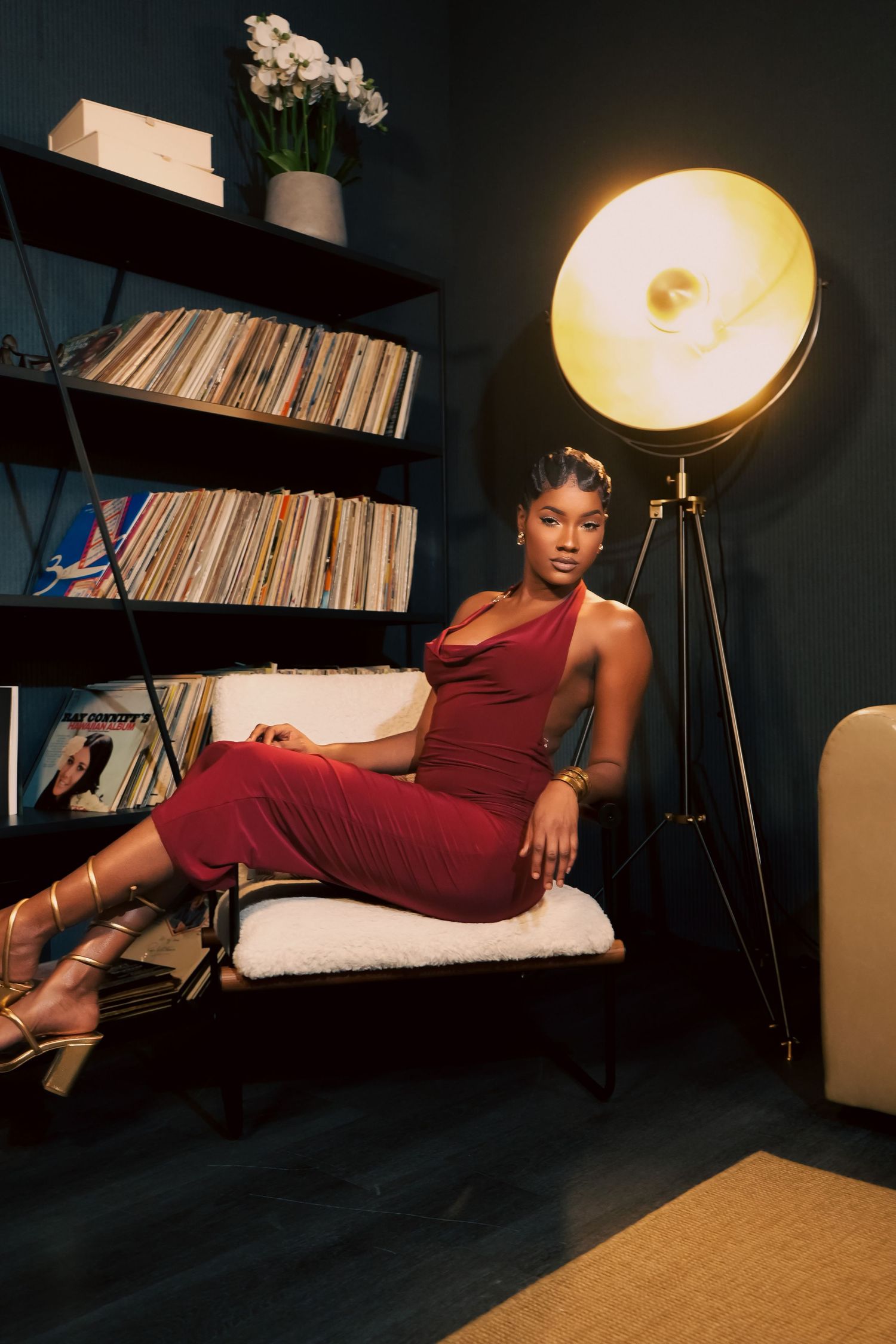 Stylish figure in burgundy dress lounges on chaise with record collection and modern lamp in moody lighting.