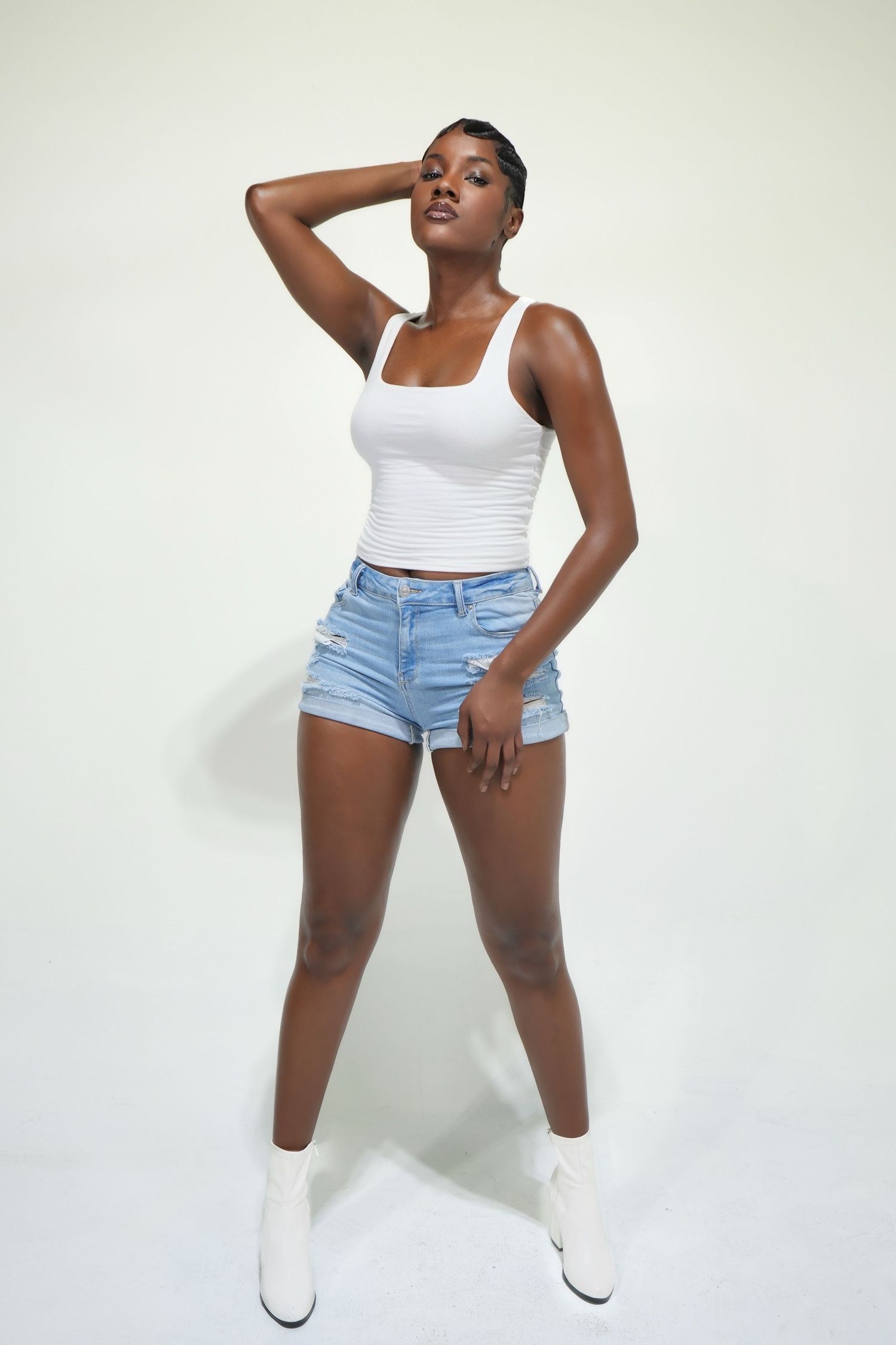 A person in a white tank top and denim shorts poses expressively against a light background in a fashion photoshoot.