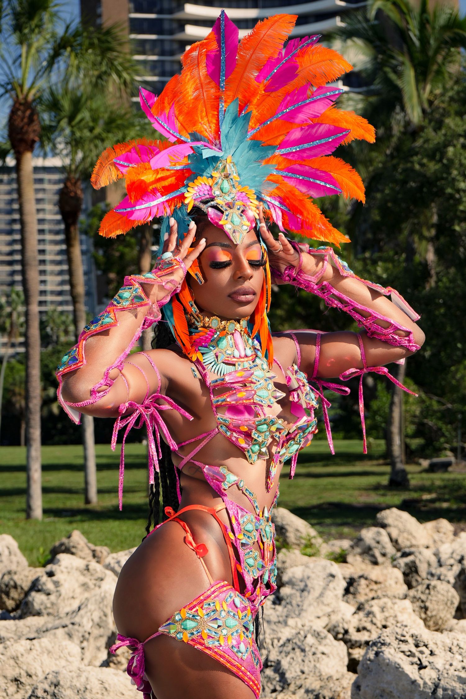 Festive carnival dancer in bright orange and pink feathered headdress performing outdoors with palm trees in background.