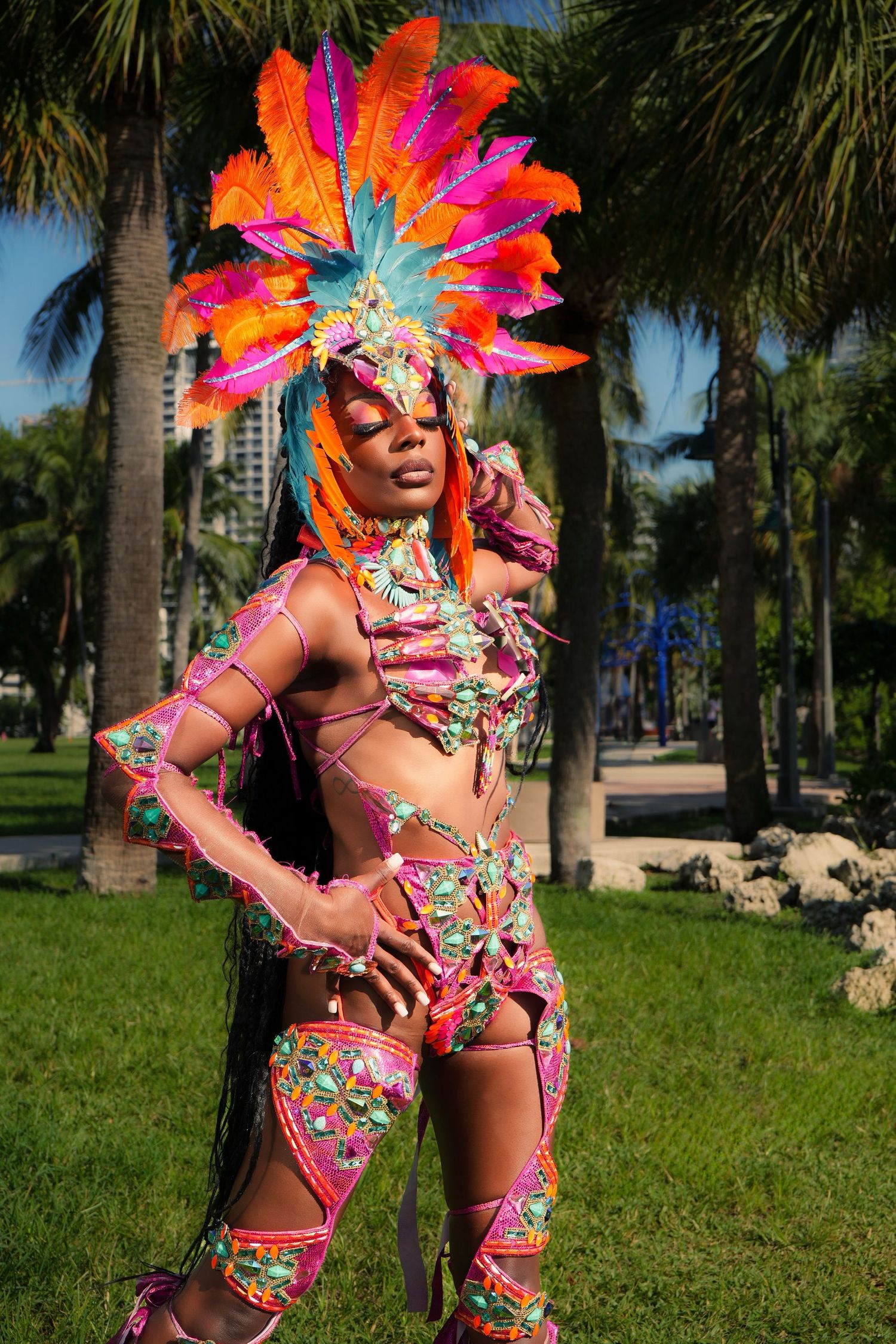 Festive carnival dancer in tropical setting wearing ornate pink floral costume with dramatic orange feathered headpiece.