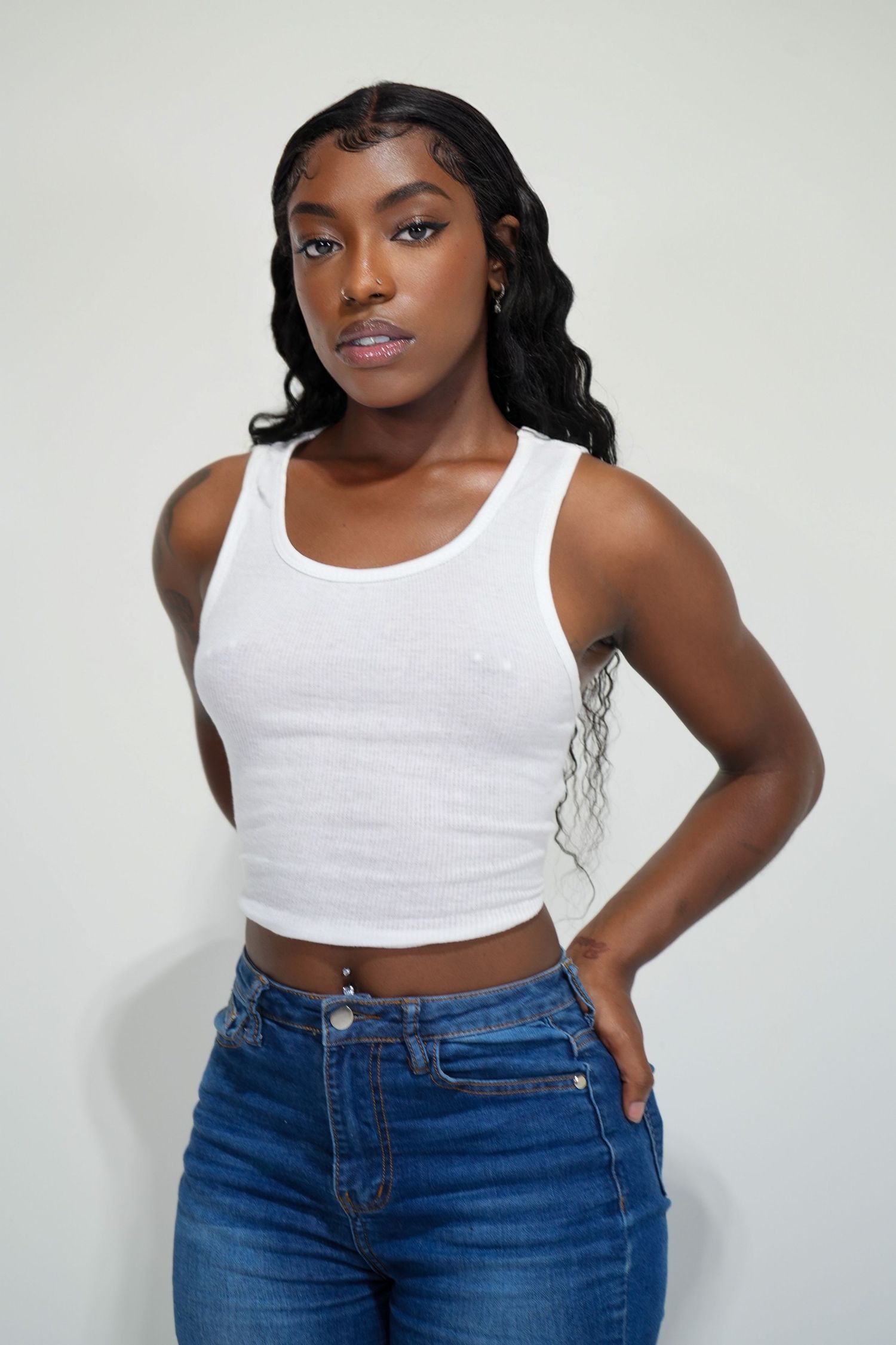 Model in white tank top and blue jeans poses against white background with wavy black hair.