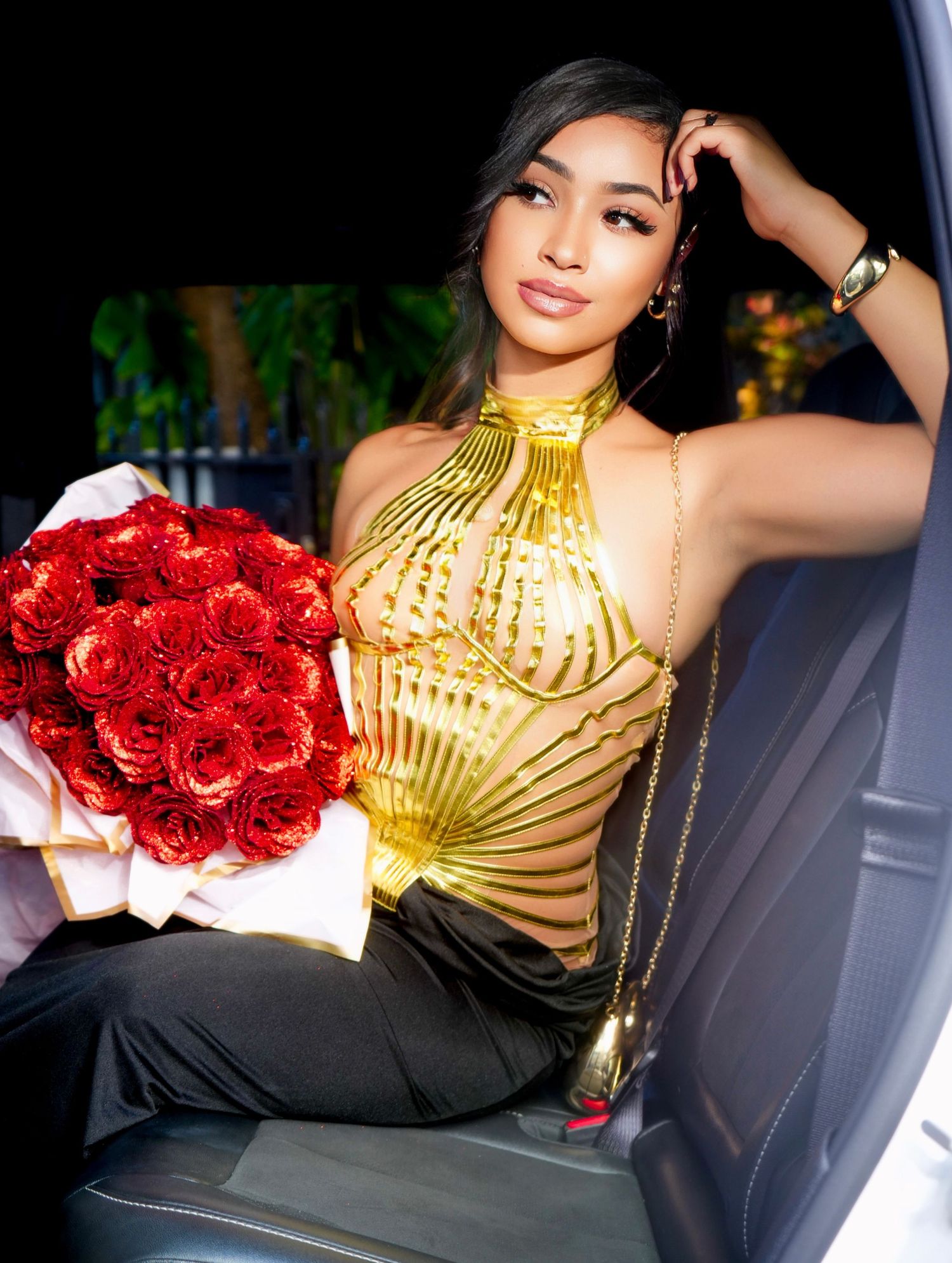 A person in a gold halter top poses in a car with a bouquet of red roses.