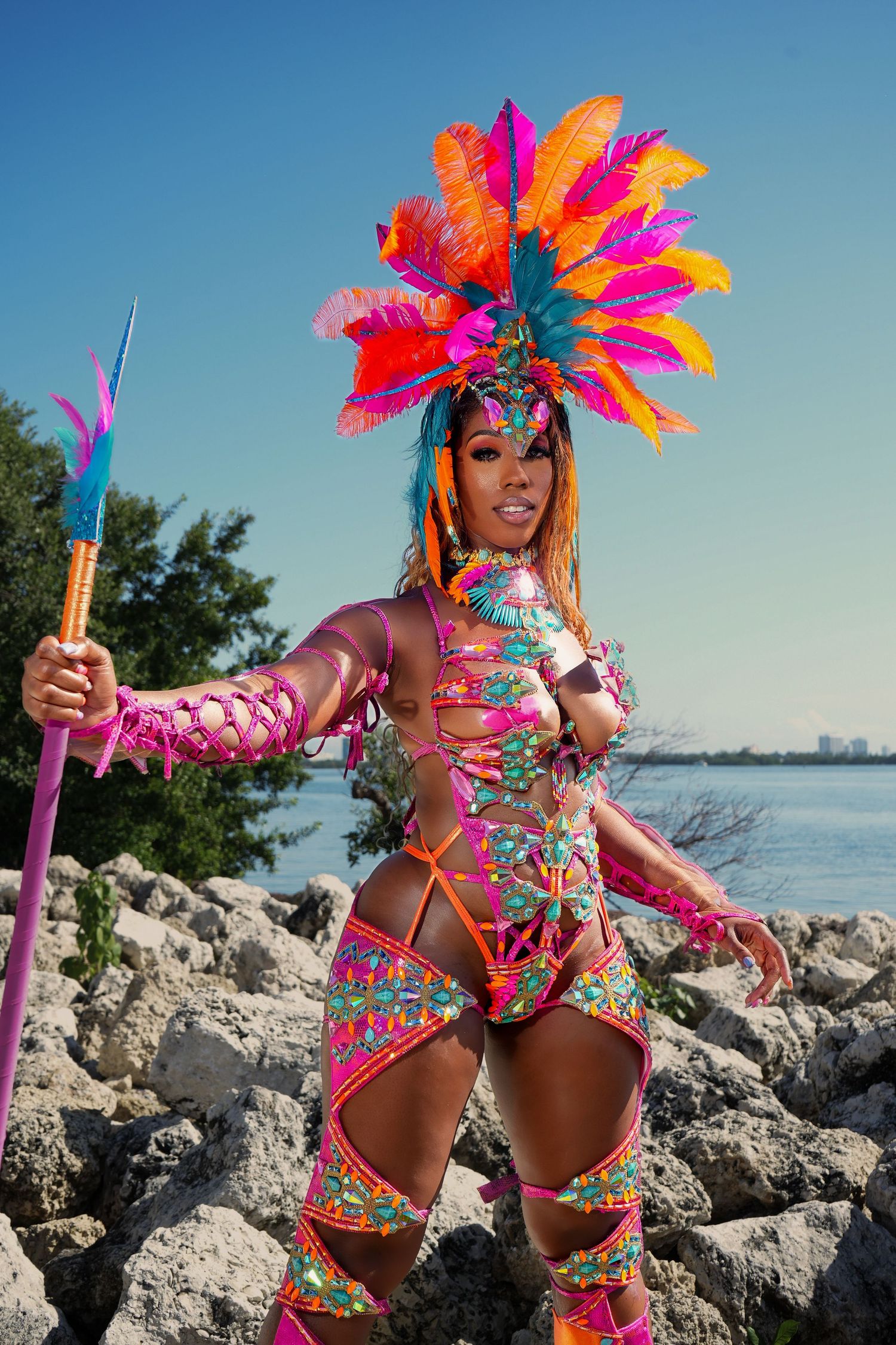 Carnival dancer in vibrant feathered headdress and colorful costume poses on rocky shoreline with ocean view.