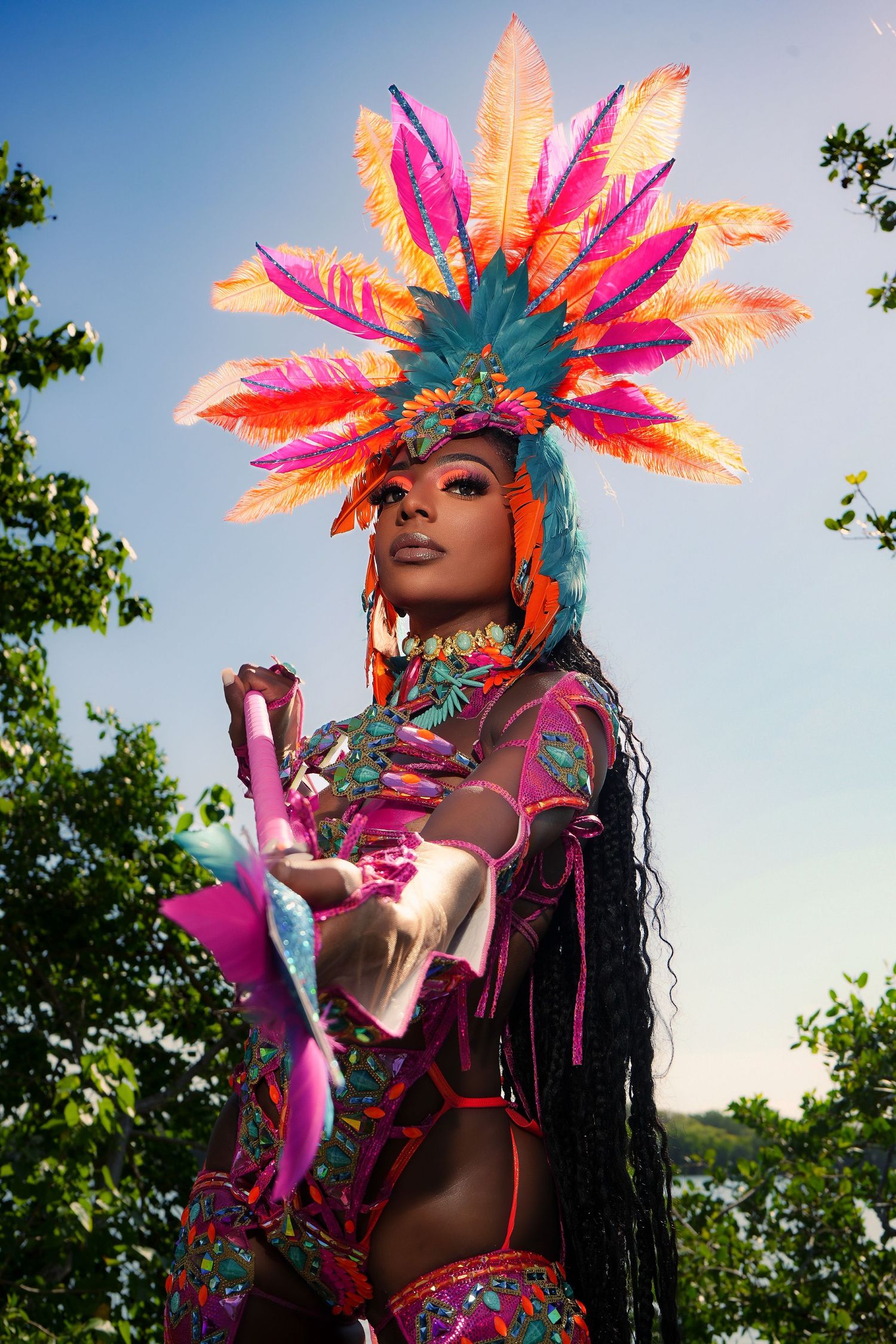 Colorful carnival performer in feathered headdress and bright costume poses with pink props against blue sky.