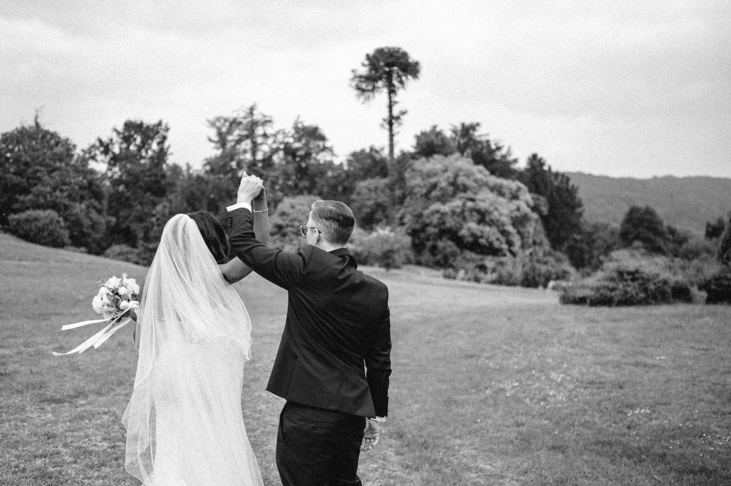 Black and white rear view of bride and groom walking triumphantly with raised hands on green lawn.