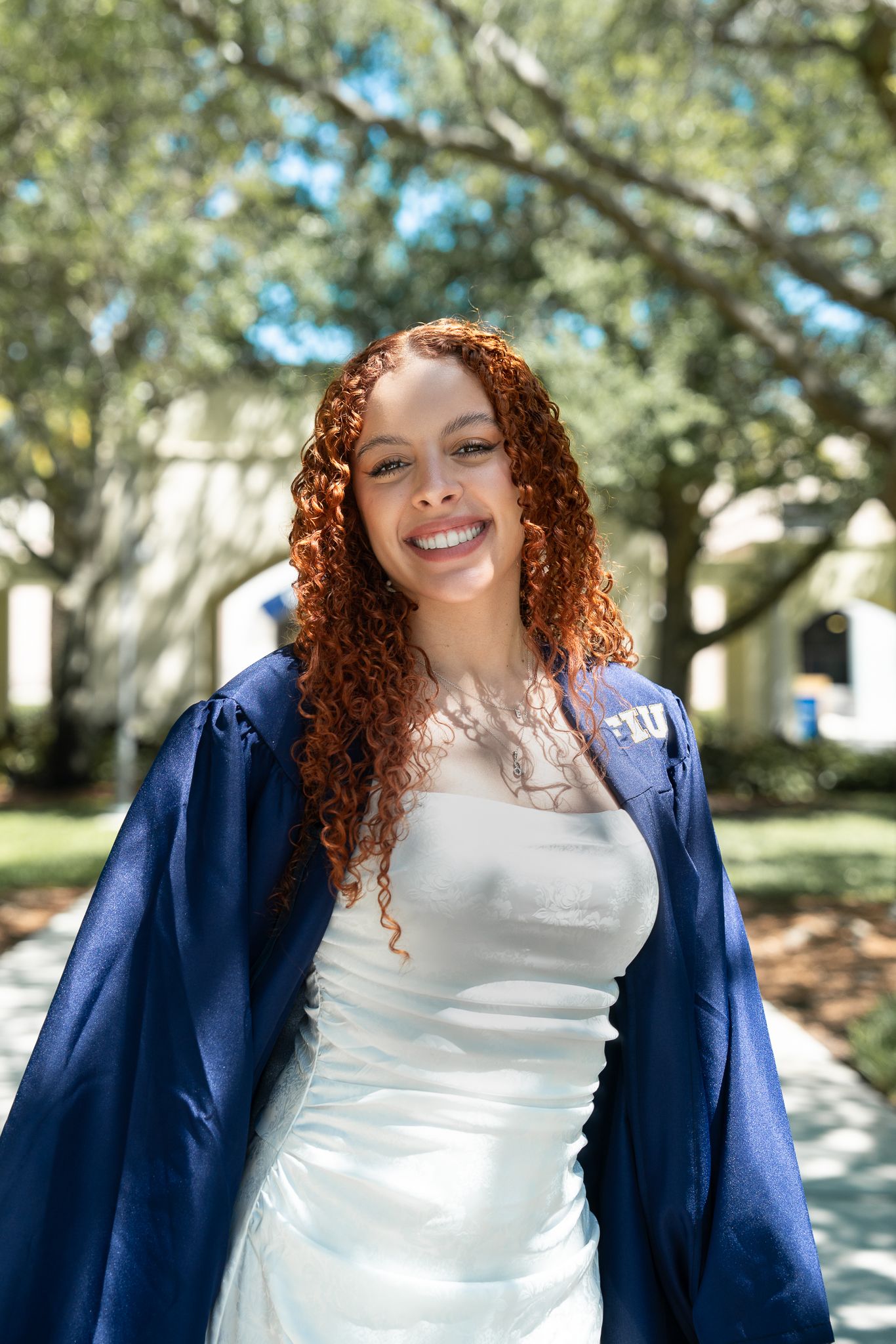 A person with curly red hair wearing a white top and navy blue blazer smiles outdoors on a sunny day.
