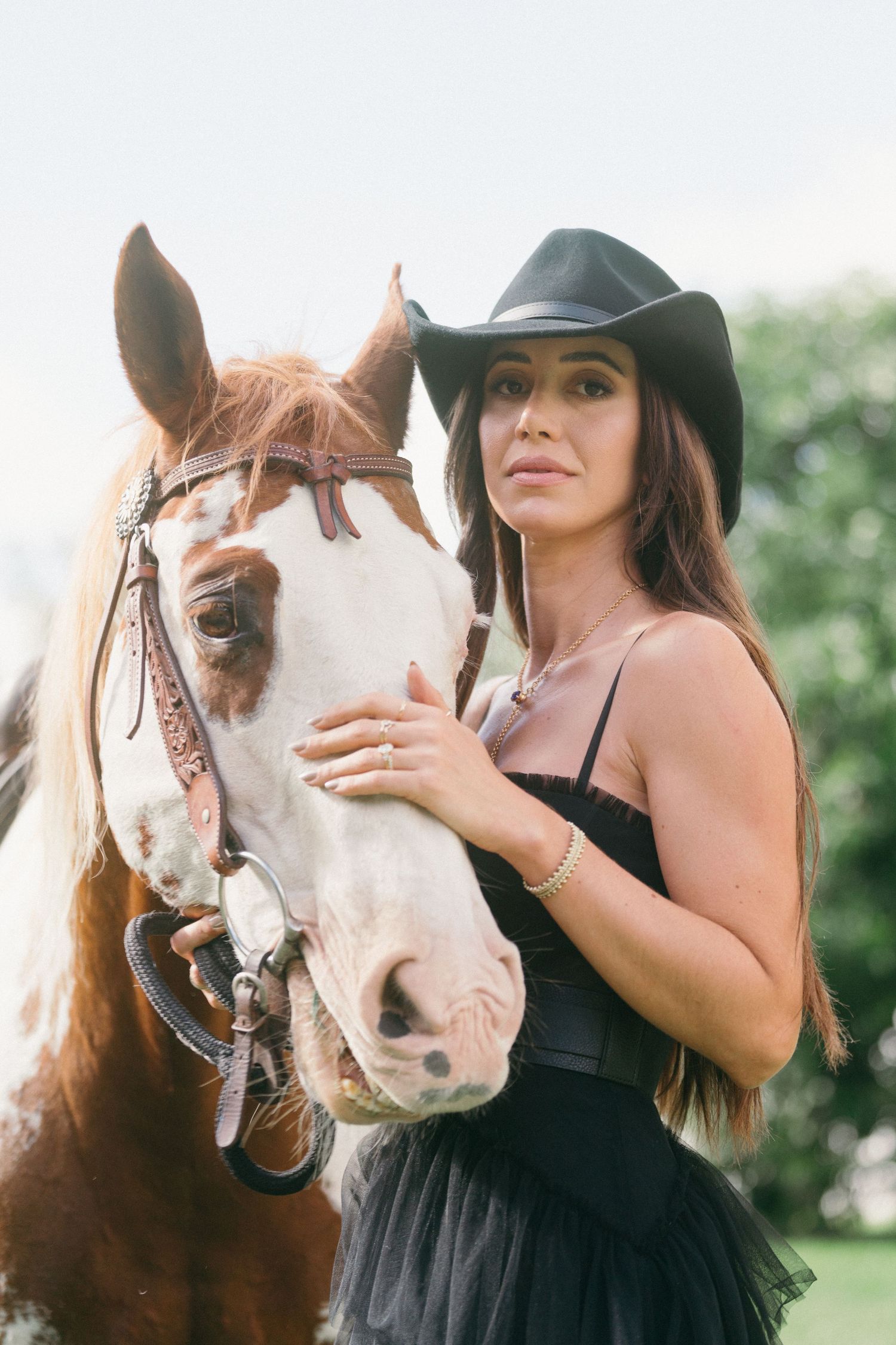 Woman in black dress and hat poses with brown and white horses in garden setting.