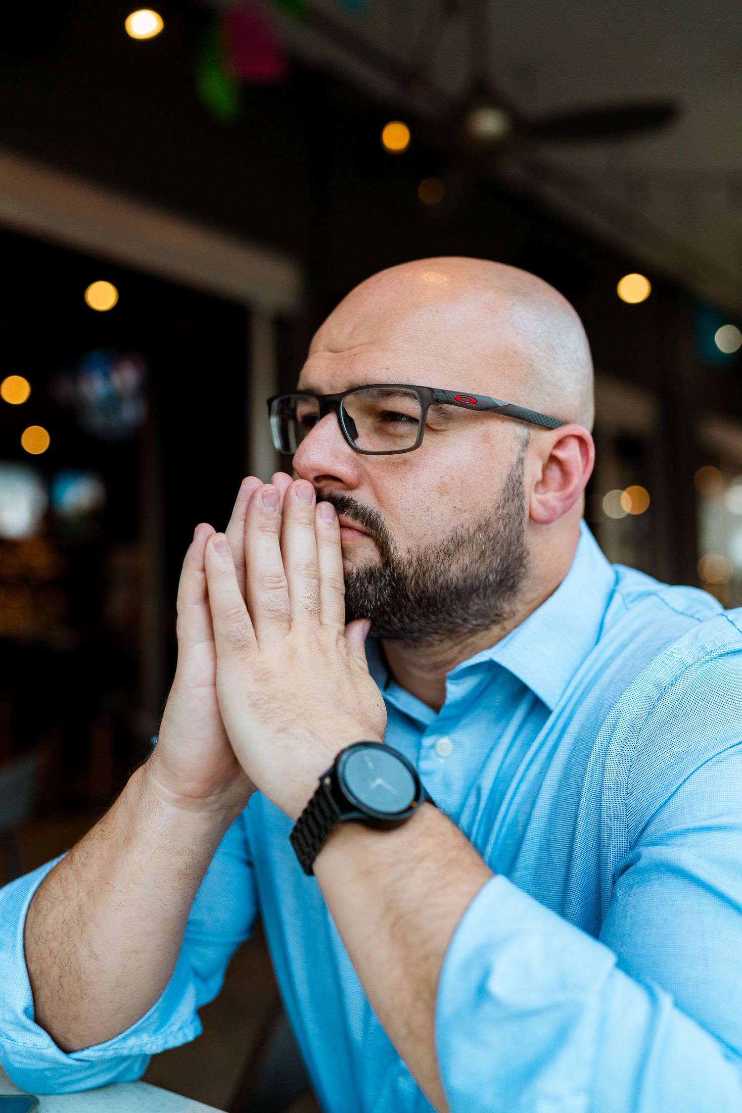 Man in light blue shirt and glasses rests his face in his hands while looking contemplative at a restaurant.