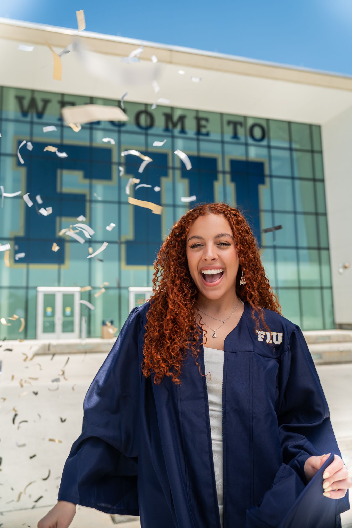 Graduate in navy blue cap and gown celebrates with confetti outside modern university building on sunny day.