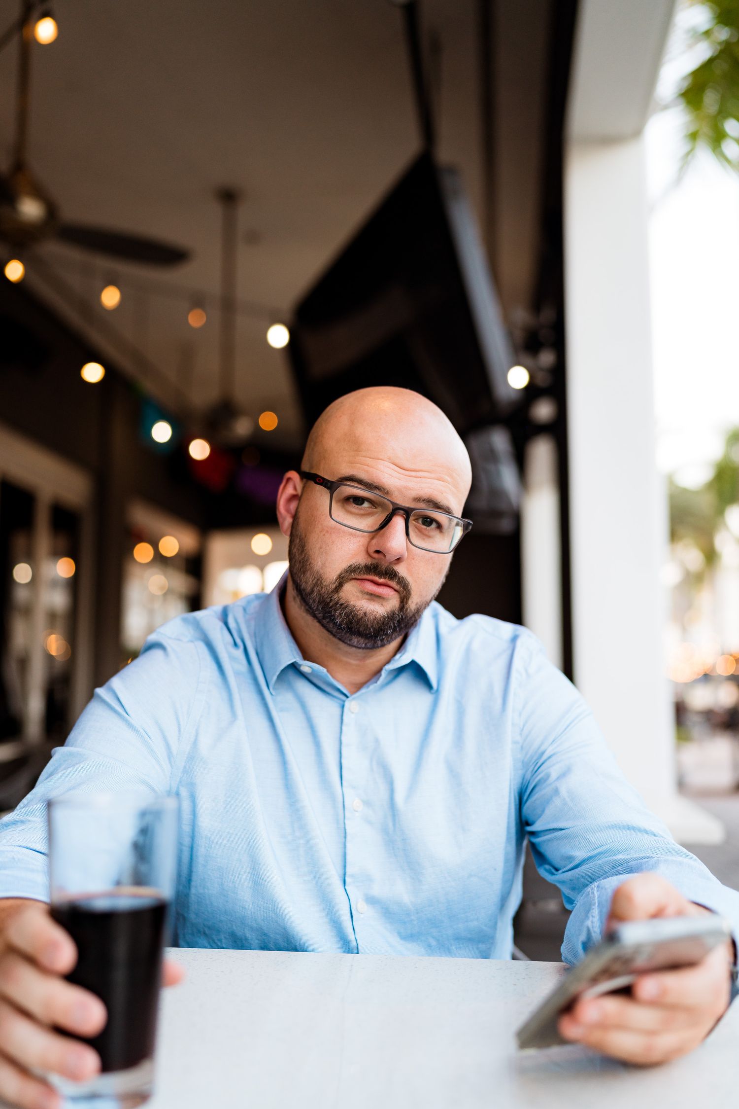 Person in light blue shirt sitting at table using mobile phone in cafe with string lights in background.