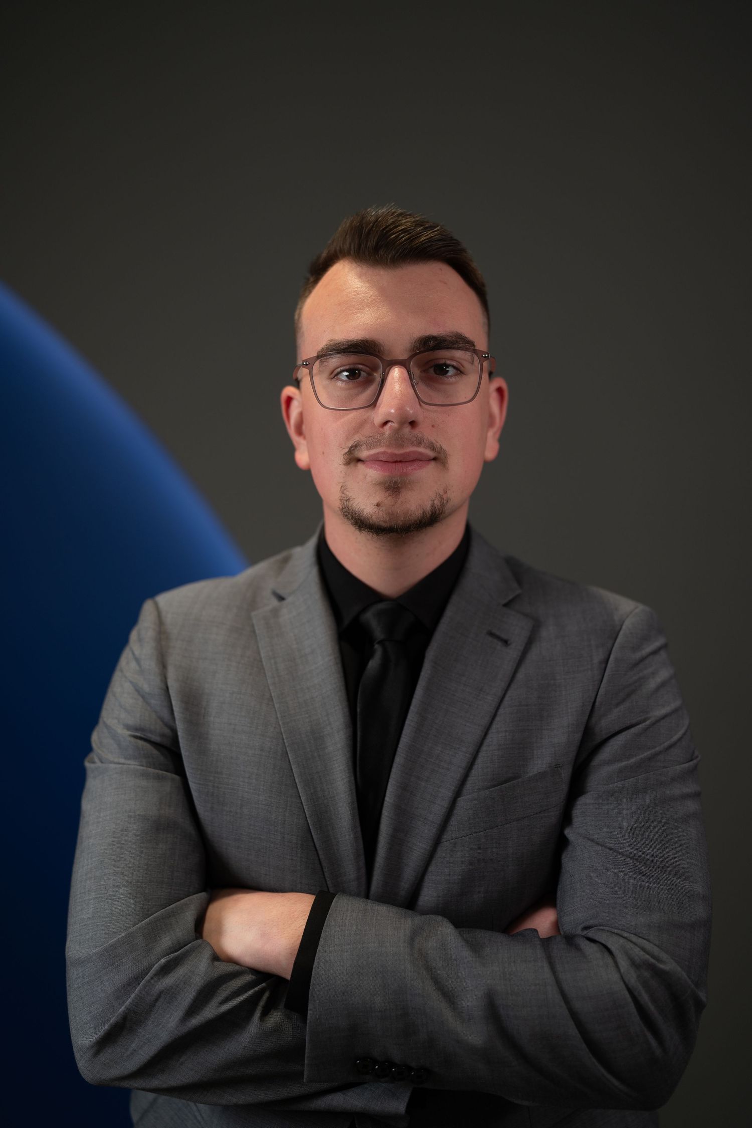 Studio portrait of person in grey blazer and black shirt against dark backdrop. heahshots