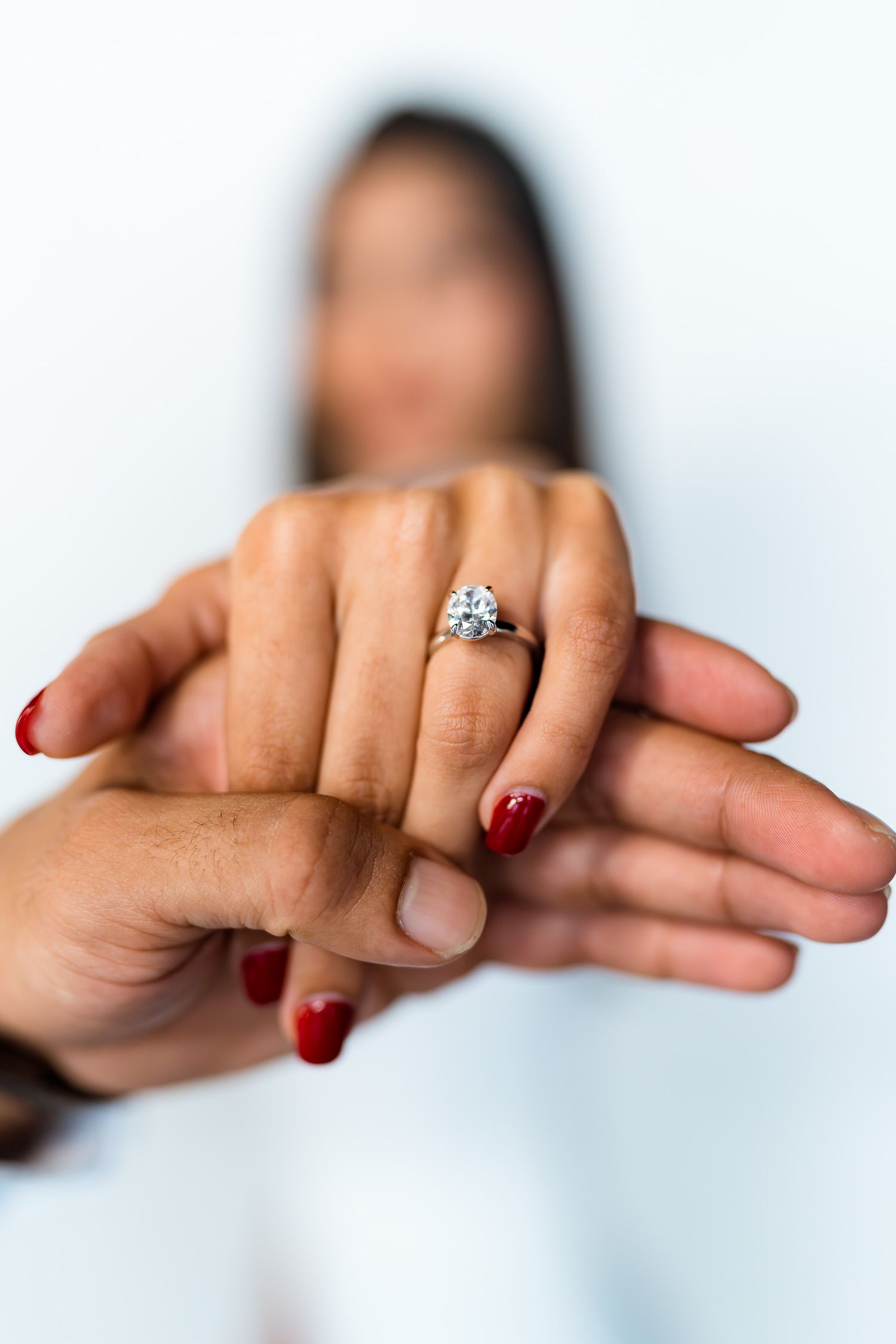 Close-up view of hands showing off an engagement ring with red nail polish against a light background.