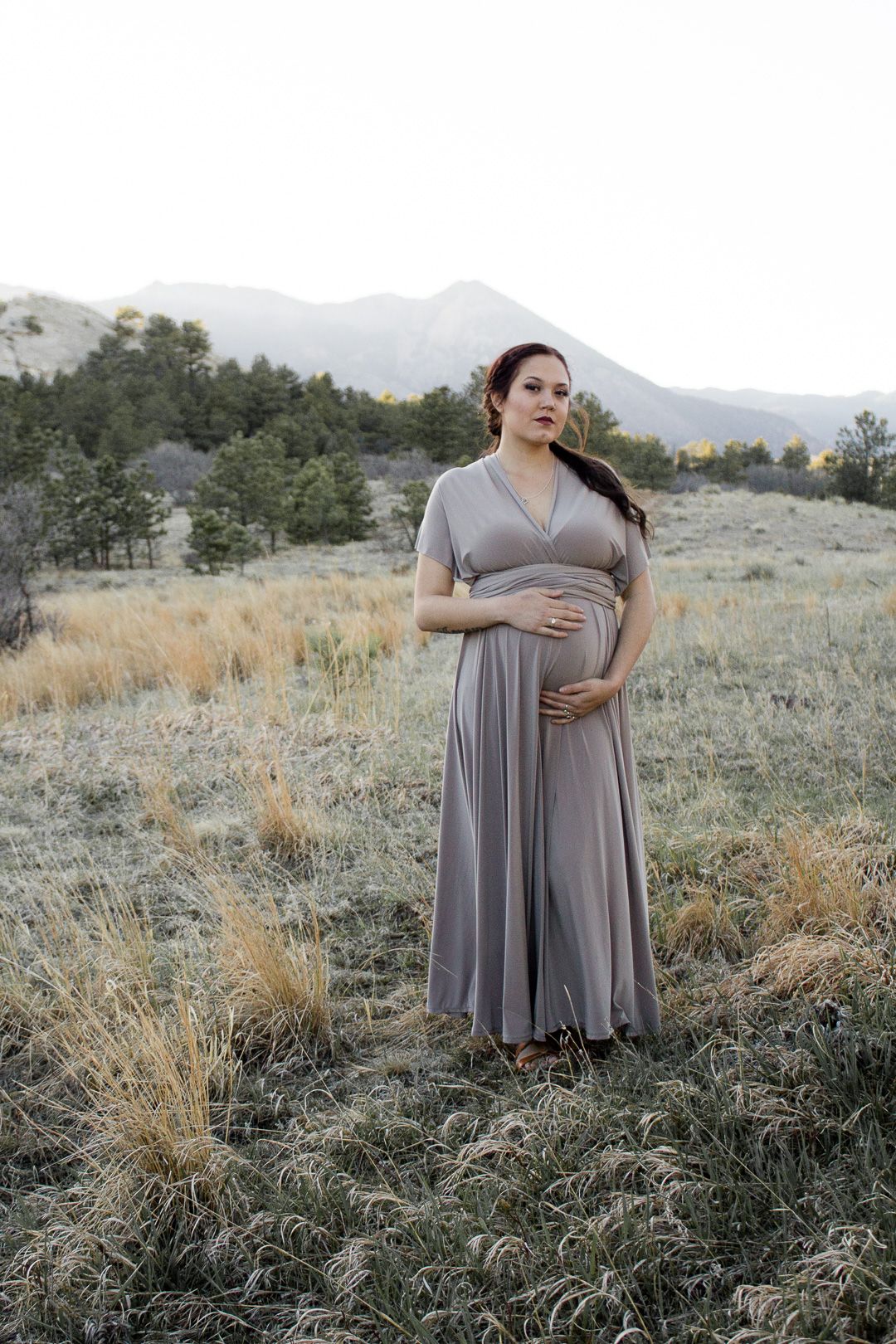 Expecting mother wearing flowy gray dress poses in mountain meadow during beautiful golden hour sunset.