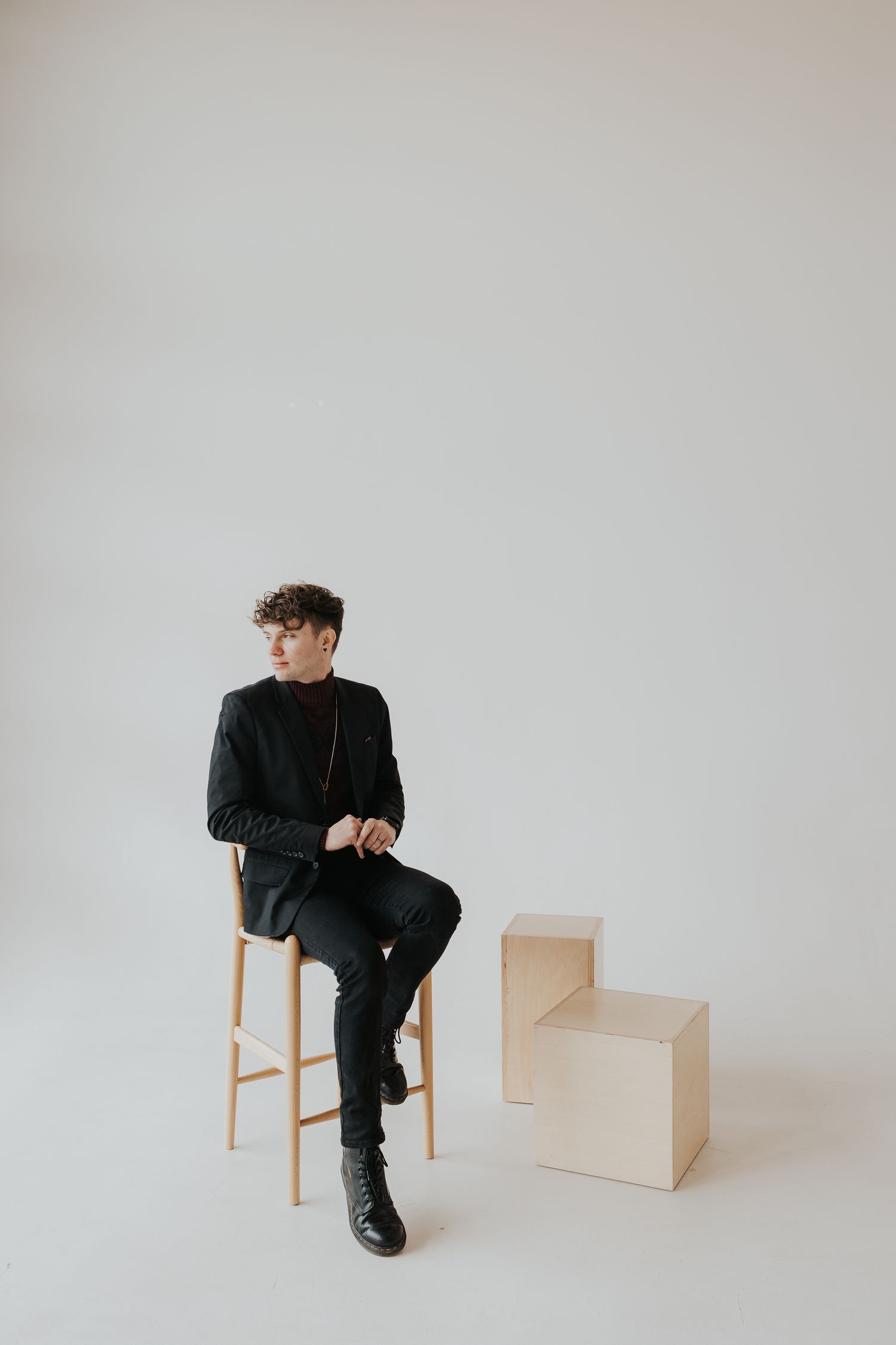 Young man in black outfit sitting on wooden stool in minimalist studio with geometric wooden blocks.
