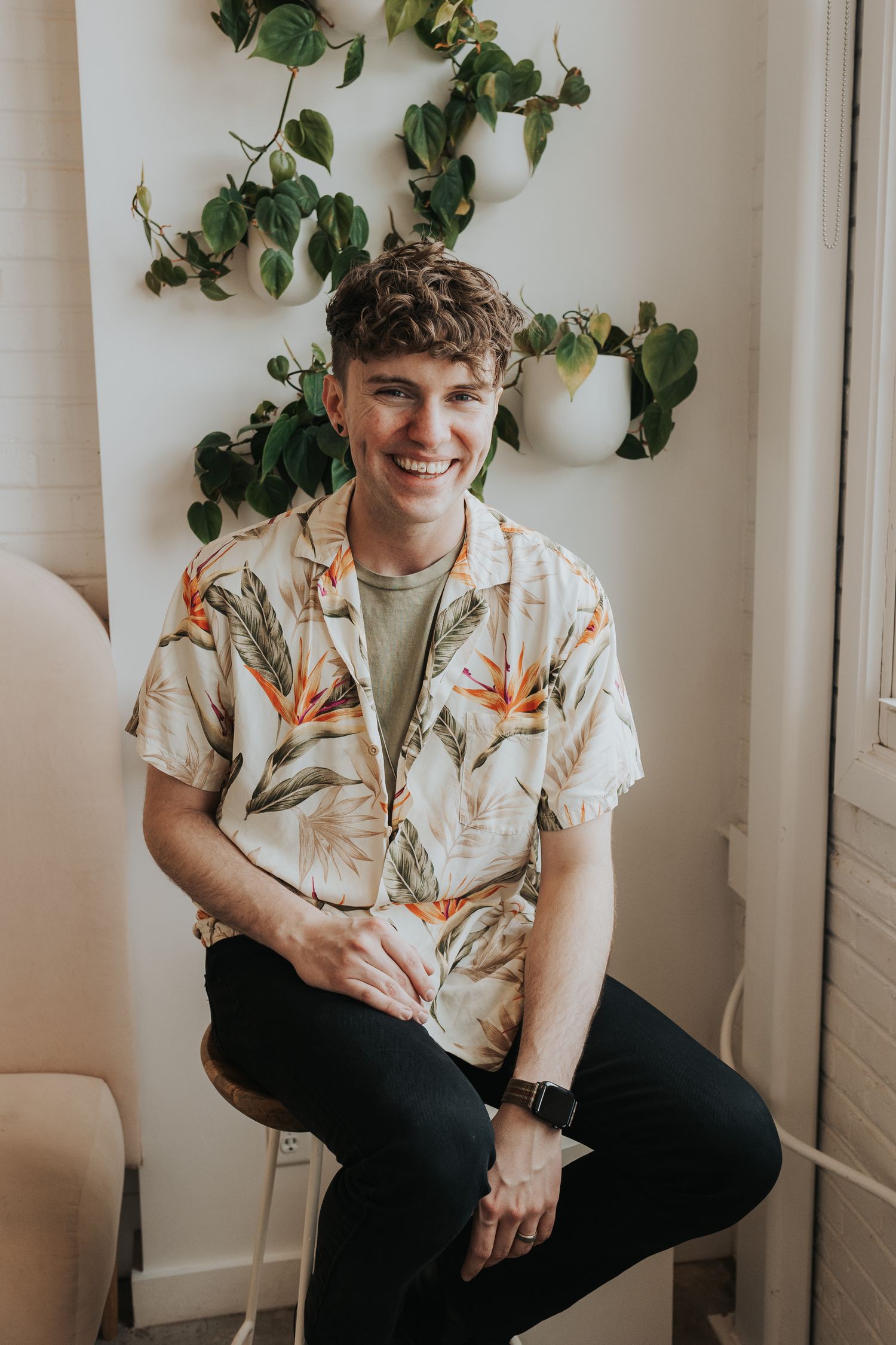 Smiling young man in floral Hawaiian shirt sitting on stool with green plants on white wall behind him.