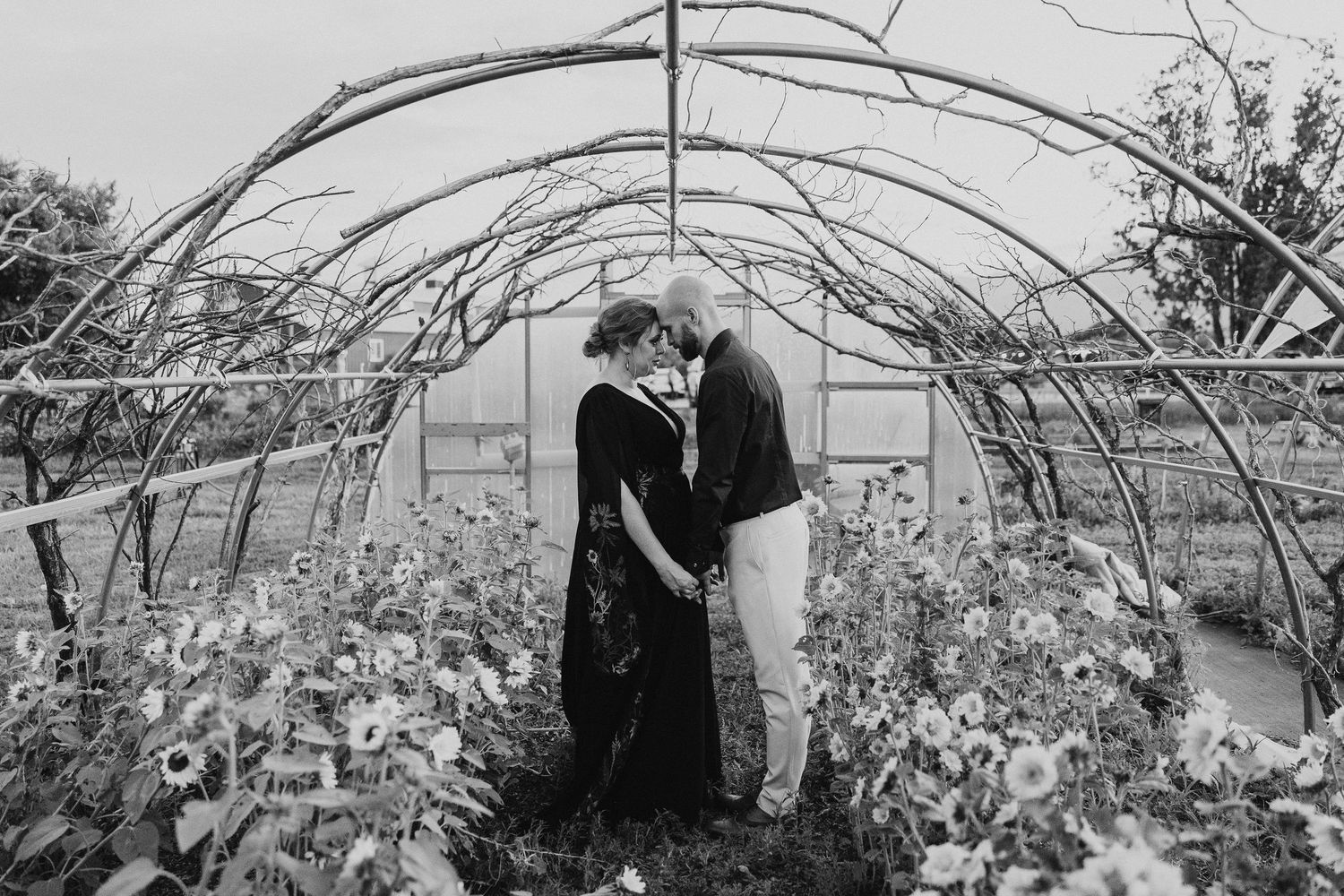Couple sharing a romantic kiss inside a flower-filled greenhouse tunnel, captured in black and white.
