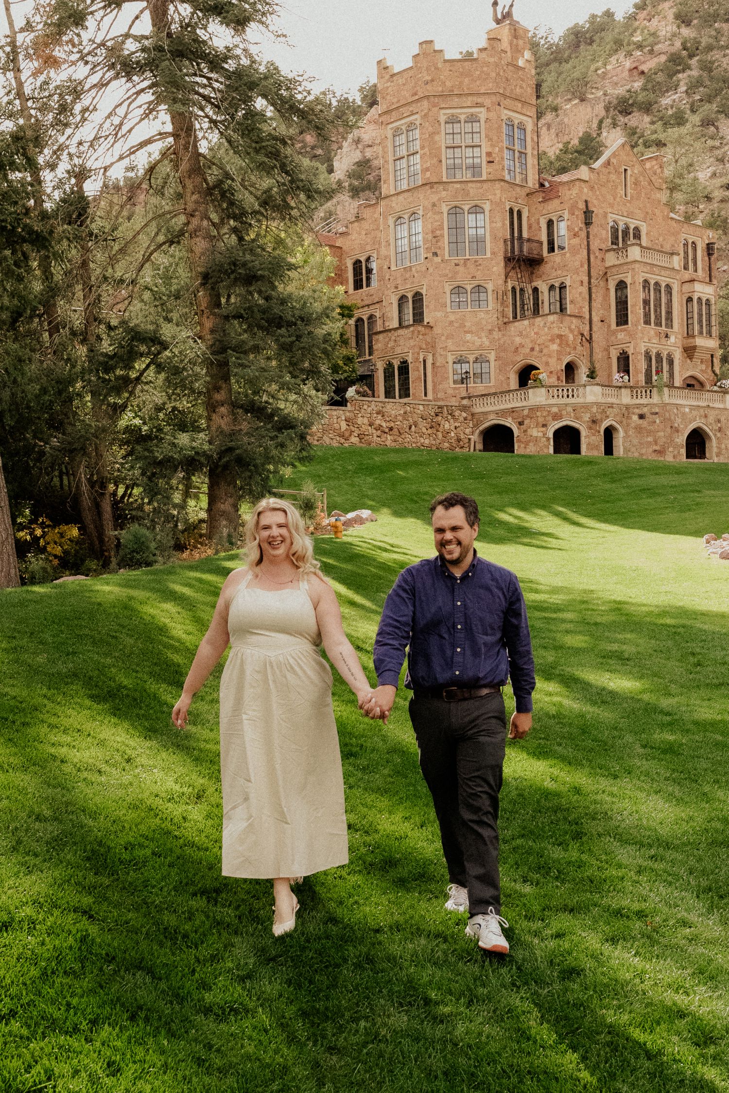 A couple holds hands walking on green grass in front of a historic stone castle building.